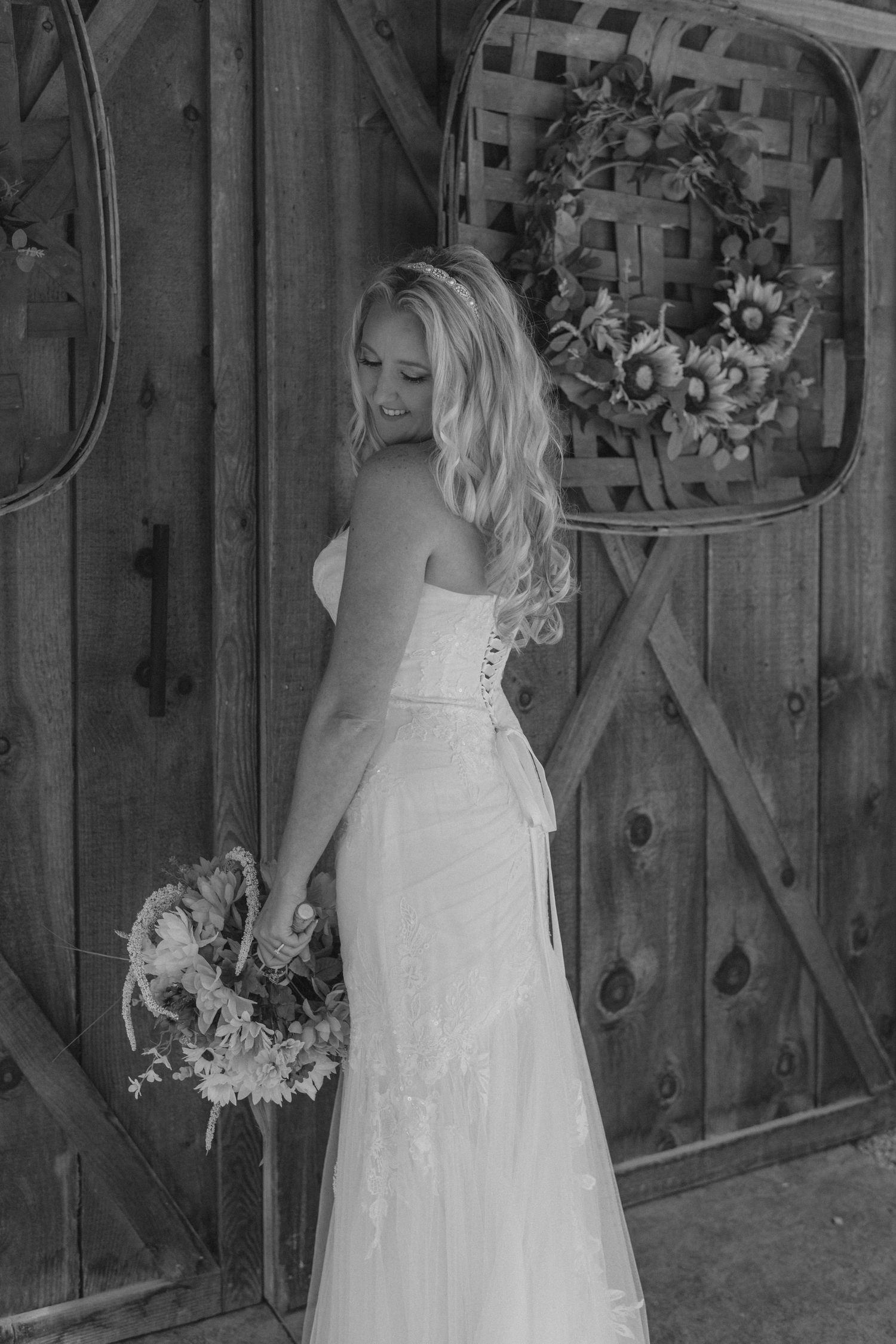 Black and white photo of bride in strapless gown holding bouquet near rustic barn wooden doors.