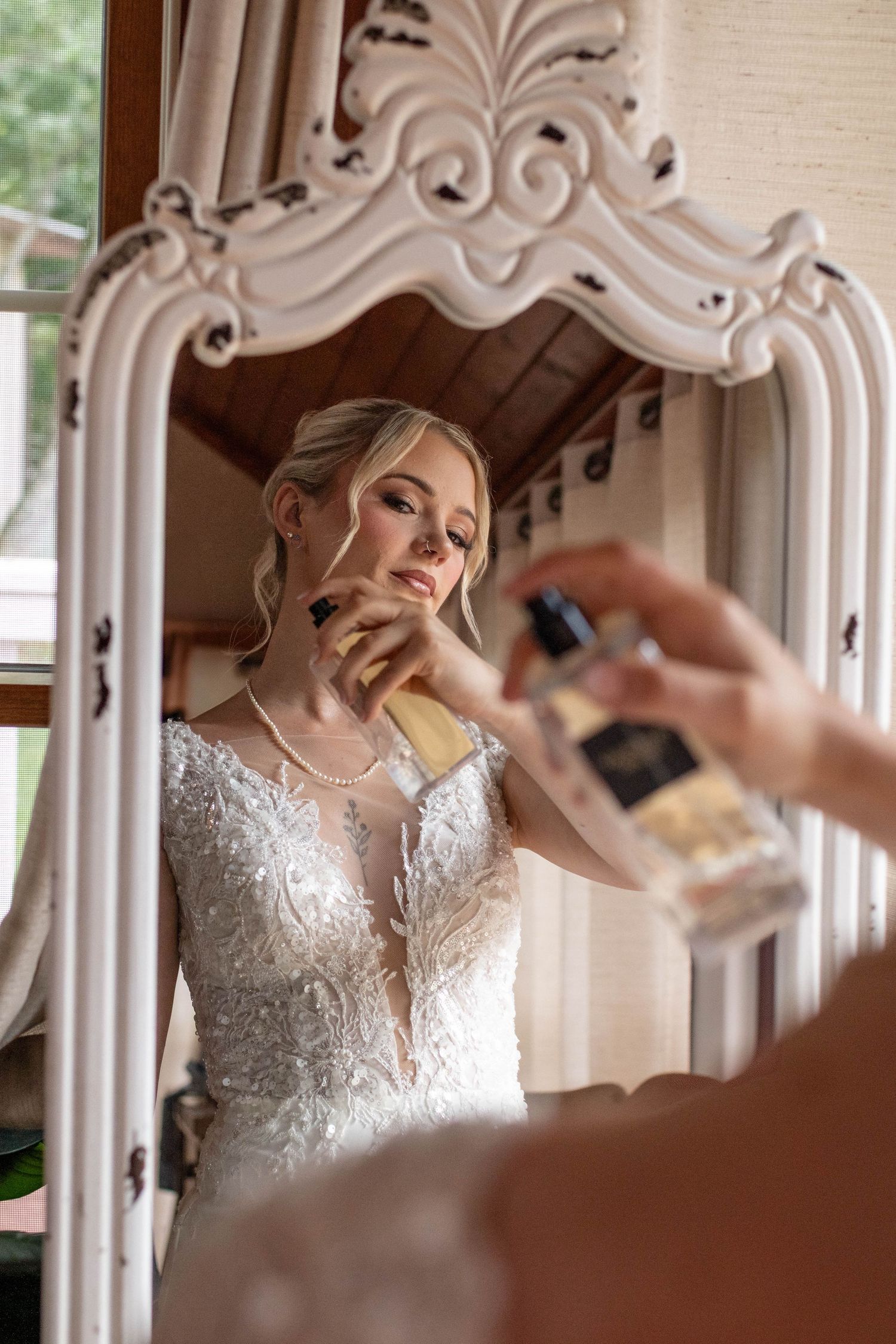 Blonde bride in lace gown applying perfume while gazing into ornate white mirror before wedding.