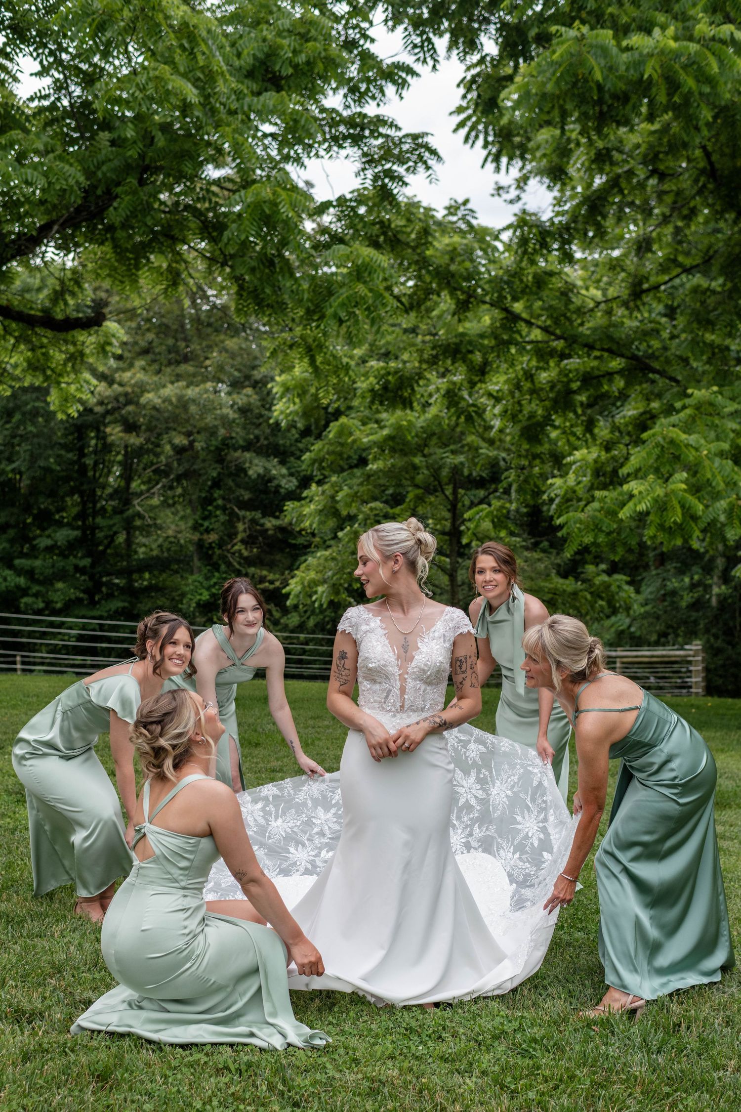 Bride in lace wedding gown surrounded by bridesmaids in sage green dresses arranging her train outdoors.