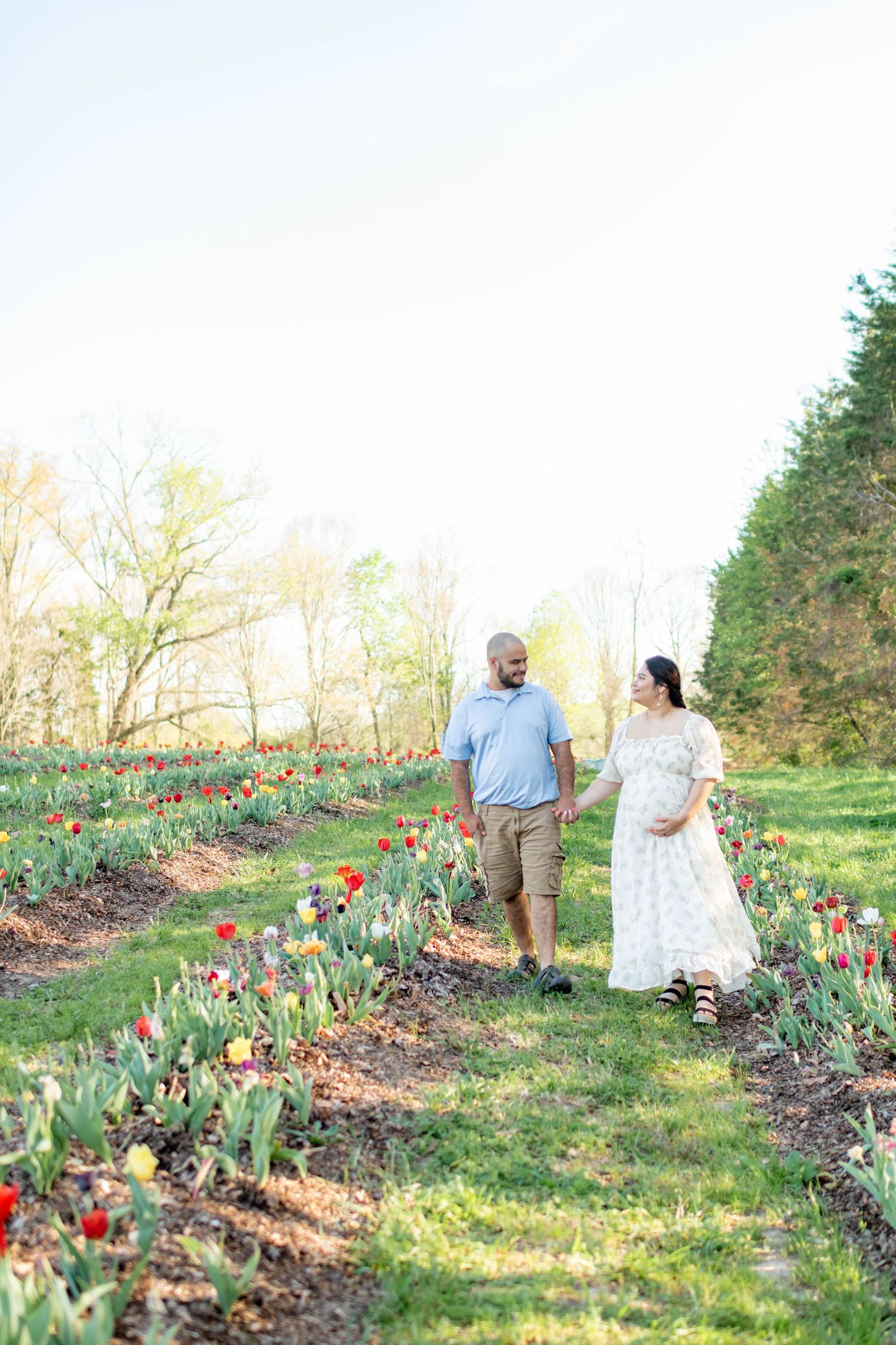 A couple walks hand-in-hand through a tulip field, the woman in a white dress cradling her baby bump on a sunny day.