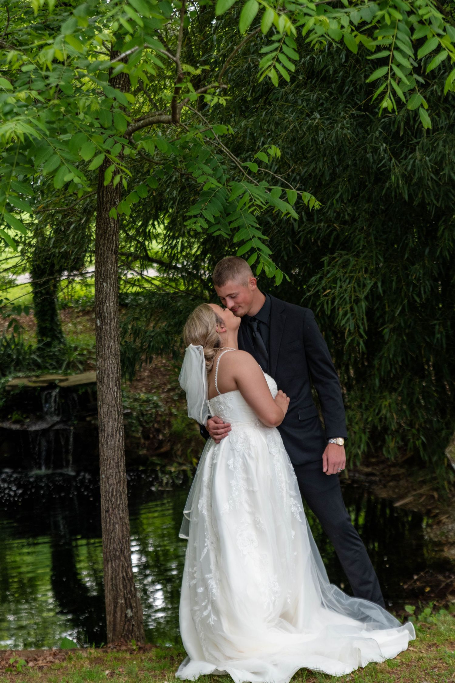 Bride and groom sharing a tender kiss near a tranquil pond surrounded by lush green trees.