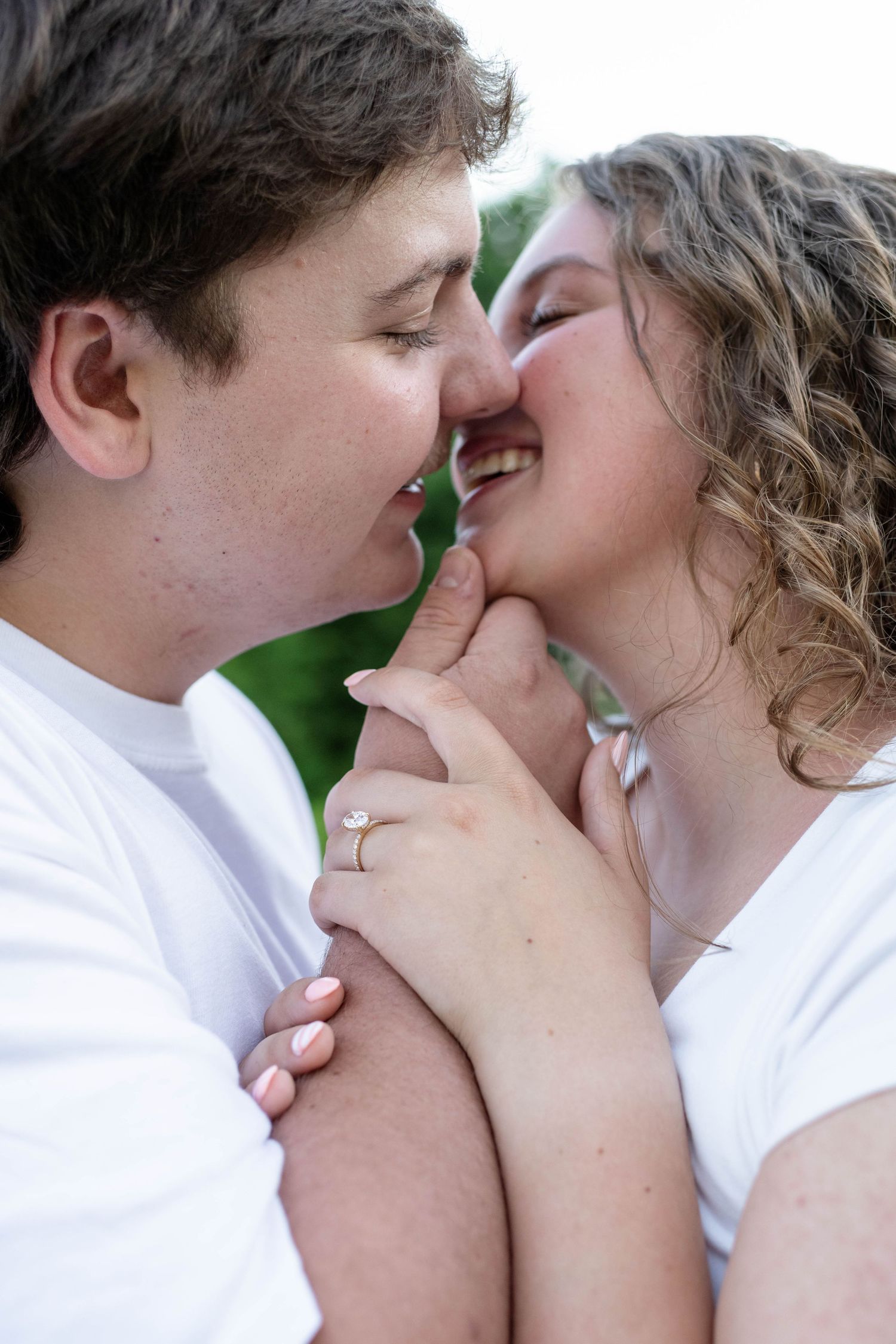 Young couple in white shirts share a tender almost-kiss while holding hands with engagement ring visible.