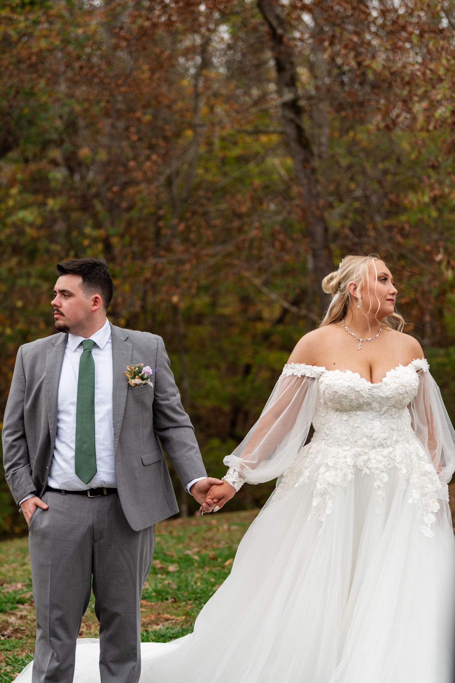 Bride in off-shoulder lace ballgown and groom in gray suit holding hands in autumn forest.