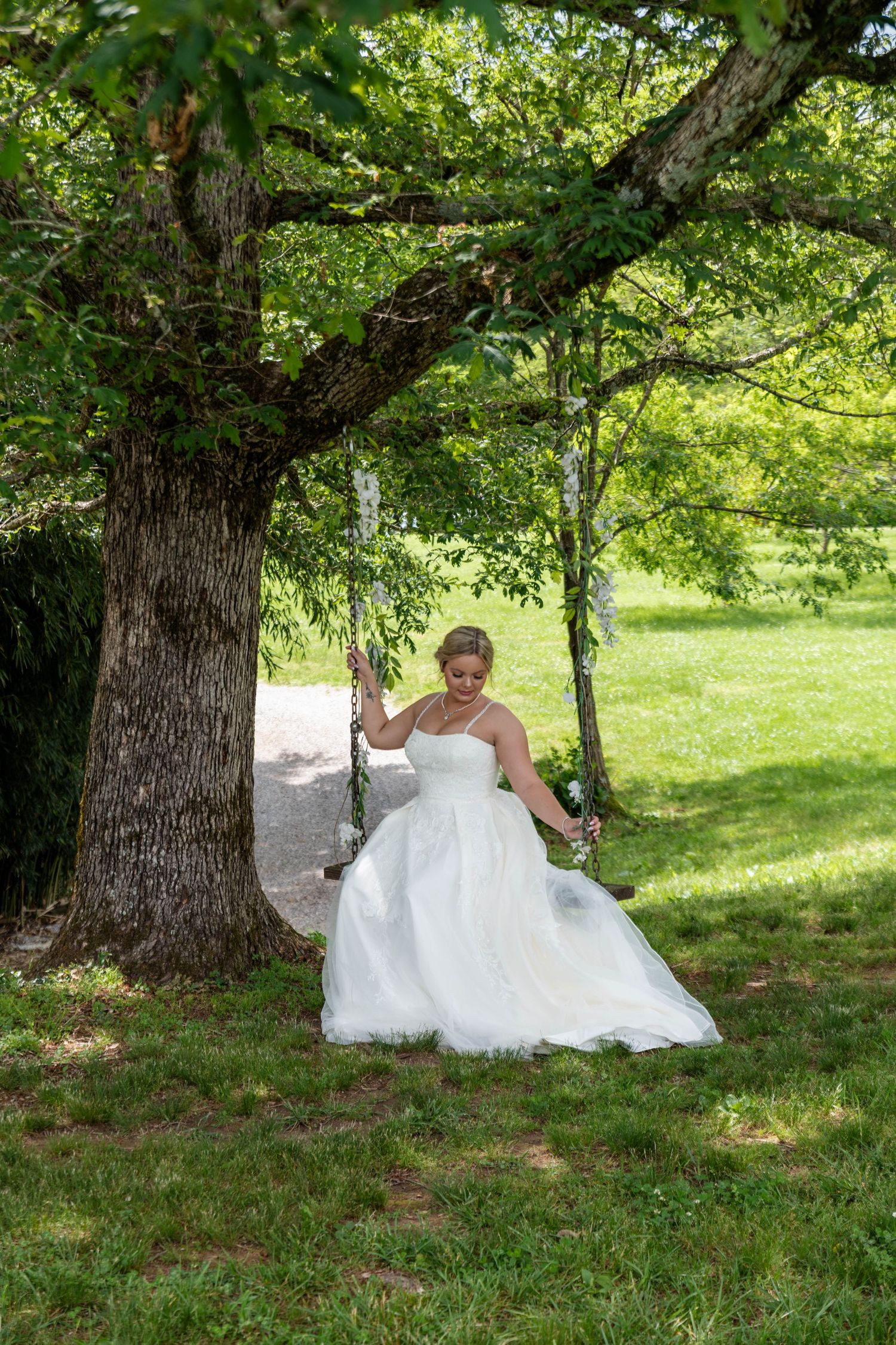 Bride in white strapless ballgown standing beside a large tree with a chain swing in sunlight.