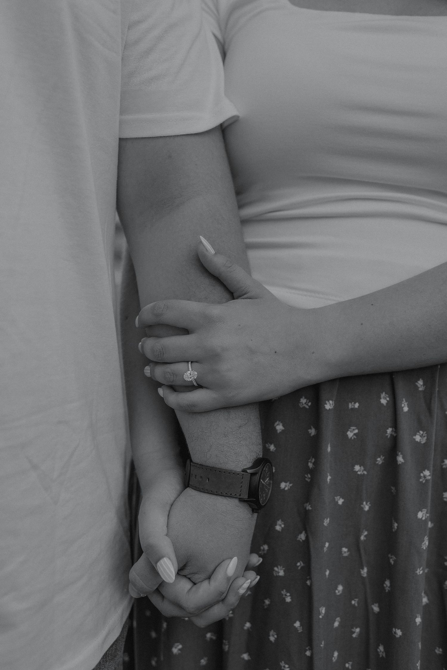 Close-up black and white photo of couple holding hands, woman wearing an engagement ring and floral skirt.