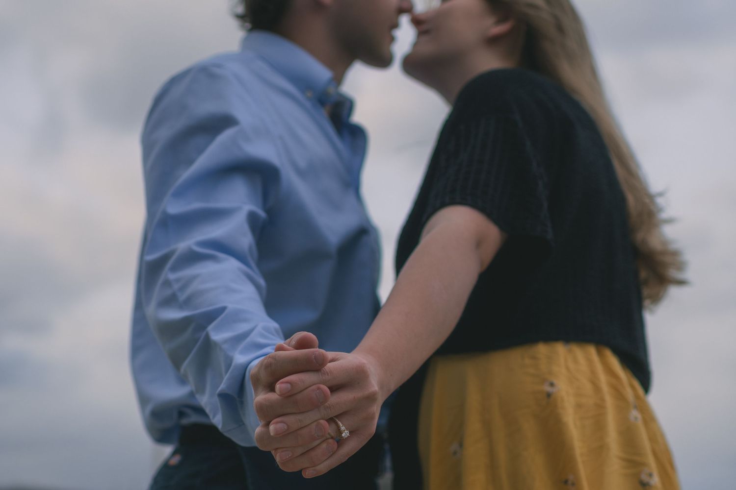 Couple holds hands and leans in for a kiss under a dramatic cloudy sky outdoors.