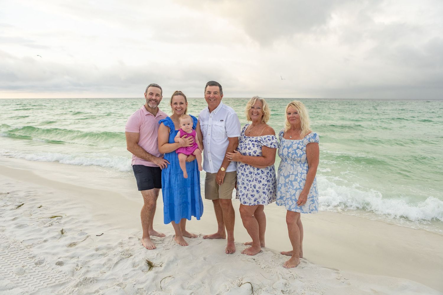 Family of five including a baby in pink poses together on a beach with crashing waves behind them.