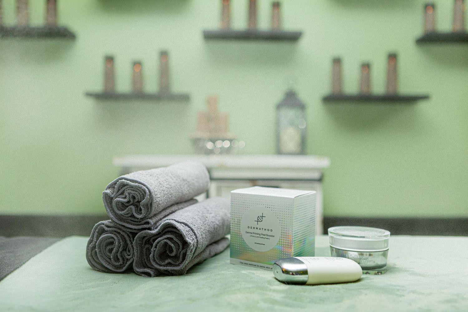Spa table with rolled gray towels, skincare products, and green walls with shelves holding candles in a serene spa setting.
