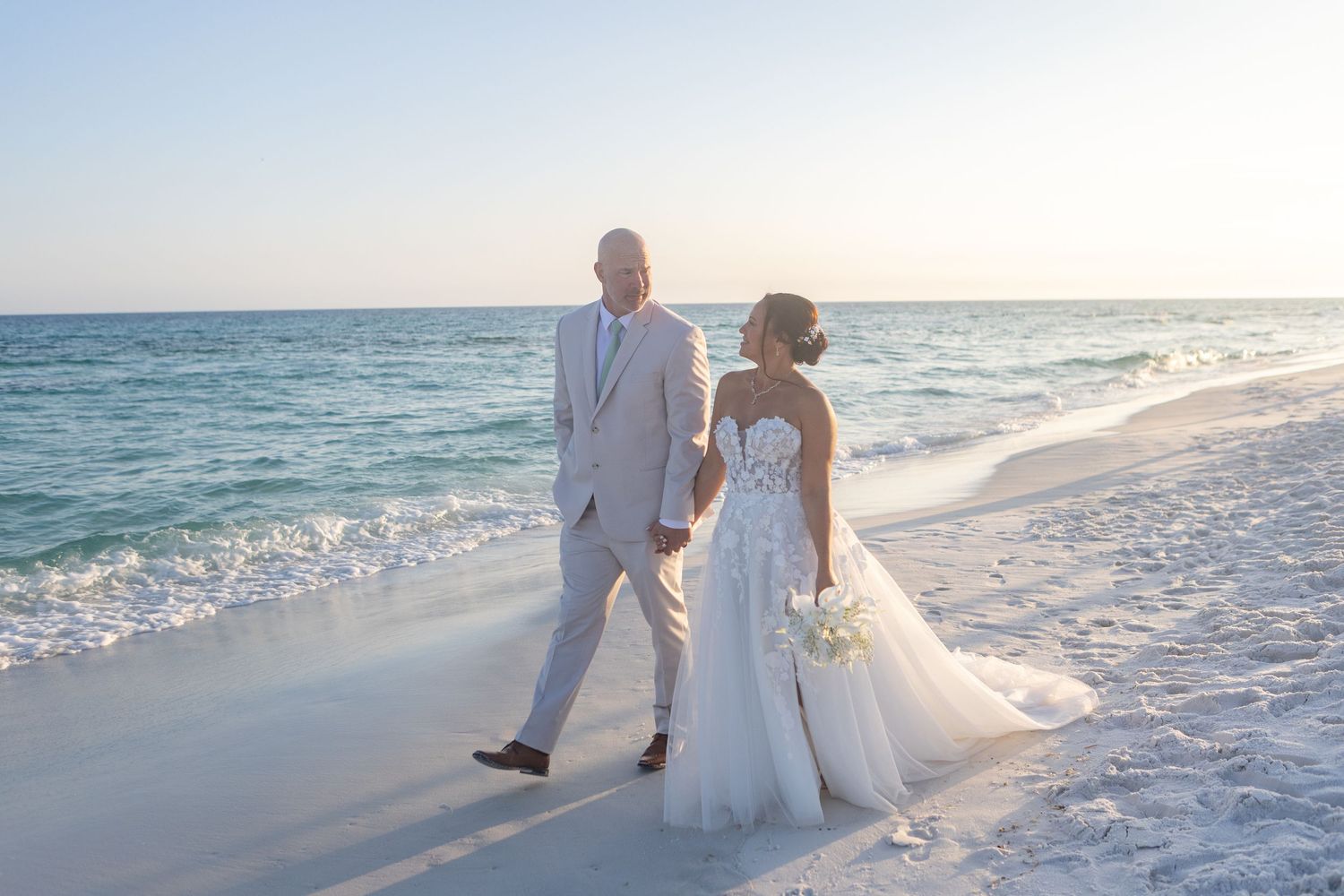 A bride and groom walk hand-in-hand along a sunlit beach, her white gown trailing in the sand.