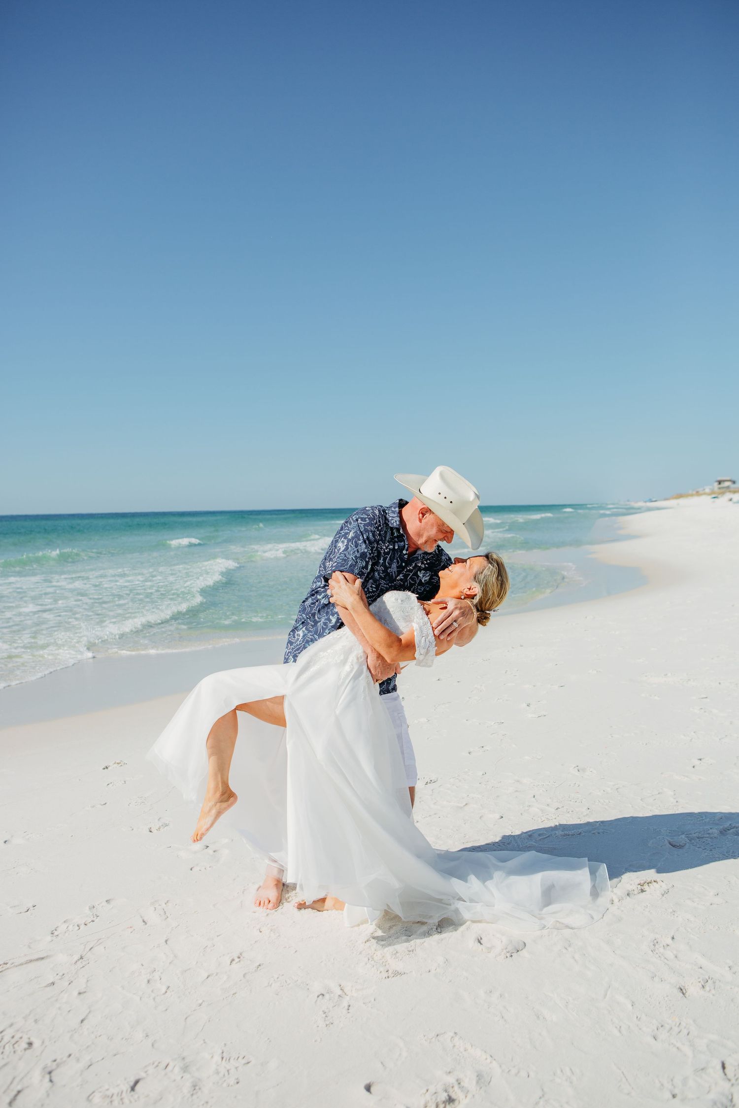 Couple sharing romantic dip kiss on white sand beach, groom in cowboy hat and navy floral shirt.