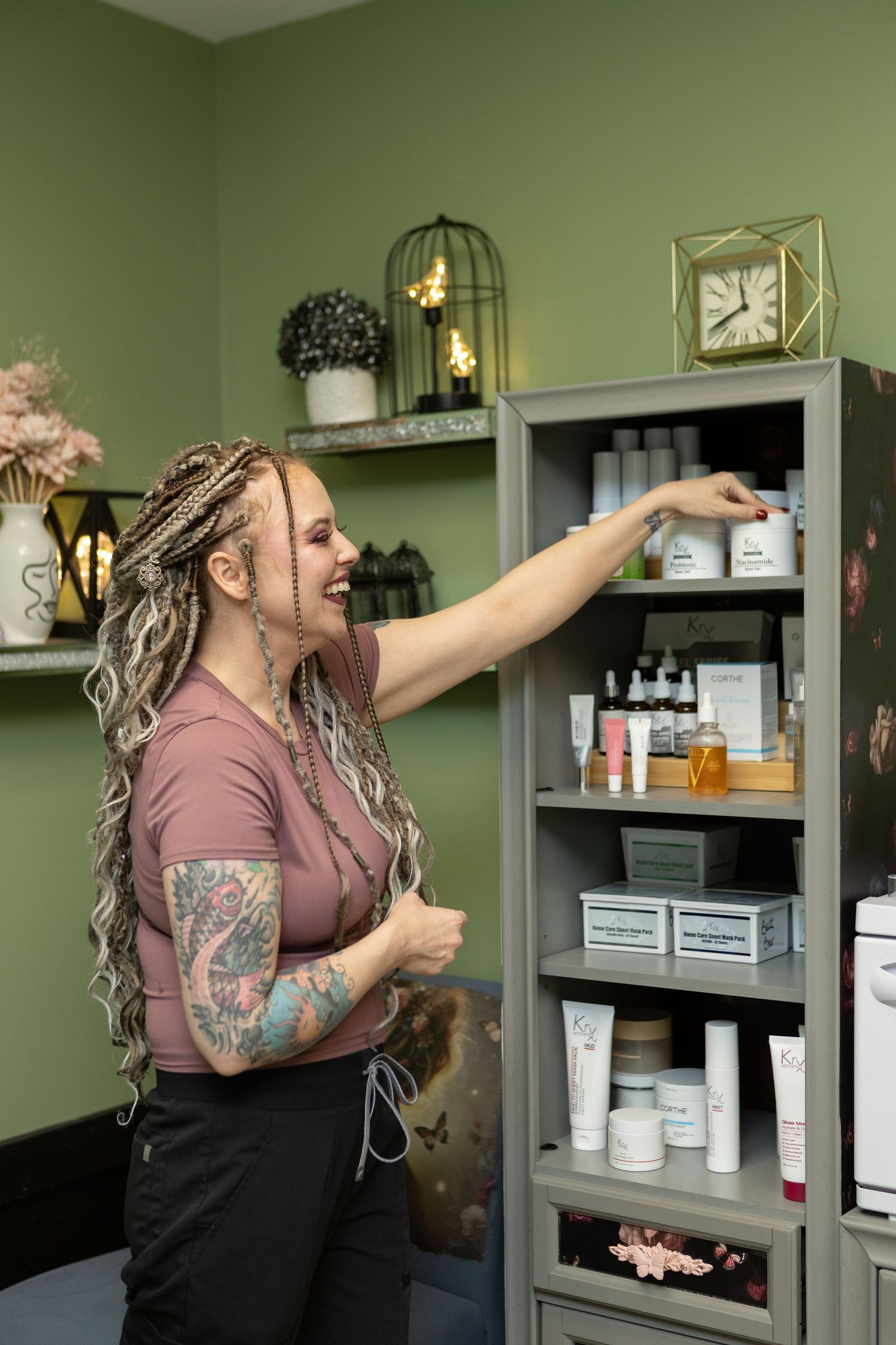 A tattooed esthetician with braided hair selects skincare products from a gray shelf in a green-walled spa.