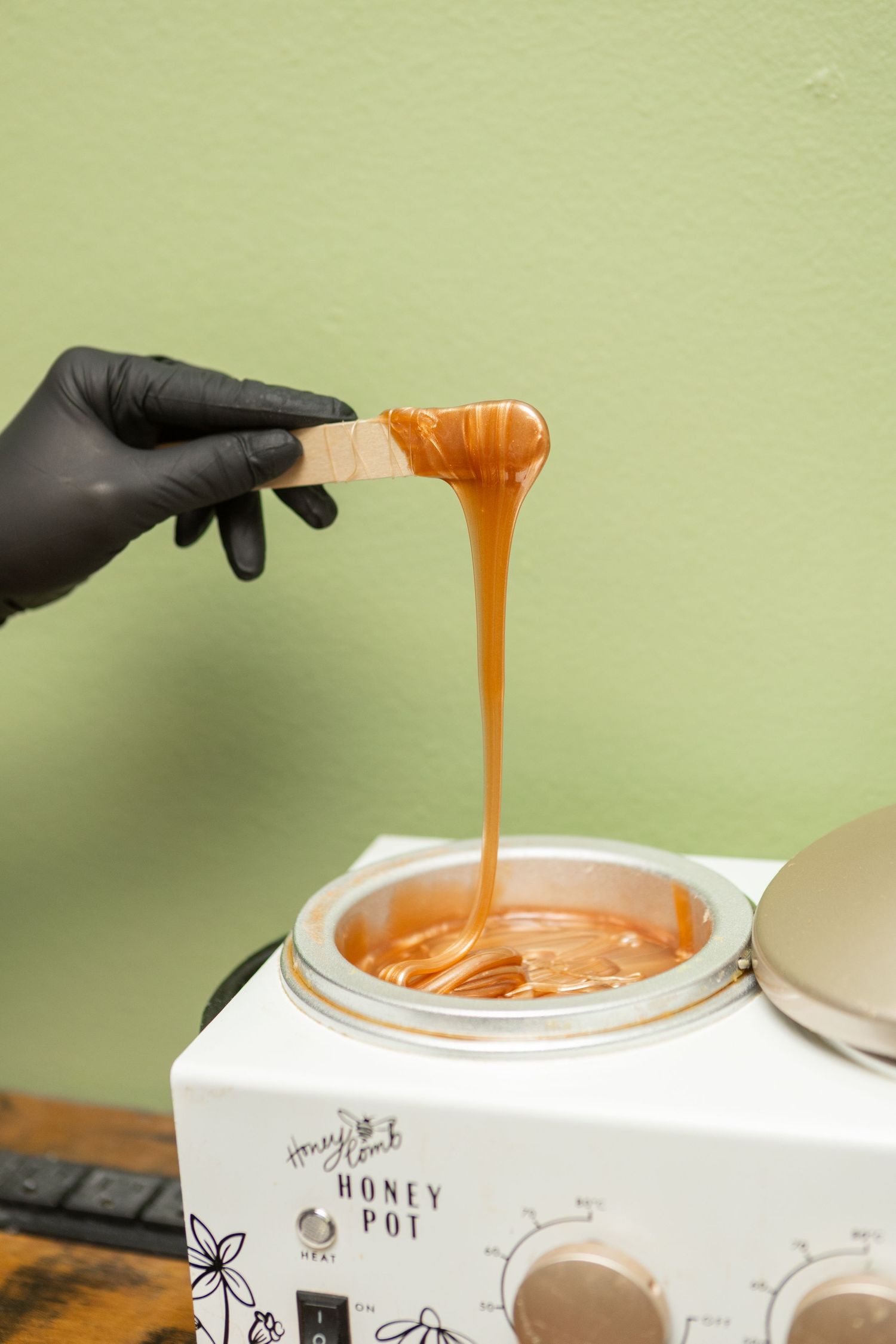 A gloved hand lifts a wooden spatula dripping warm honey wax from a Honey Pot wax warmer.