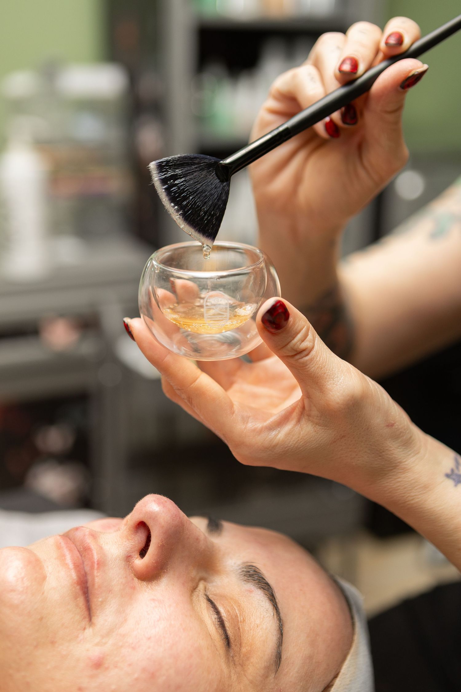 A tattooed esthetician dips a brush into a glass bowl of serum above a client during a facial.