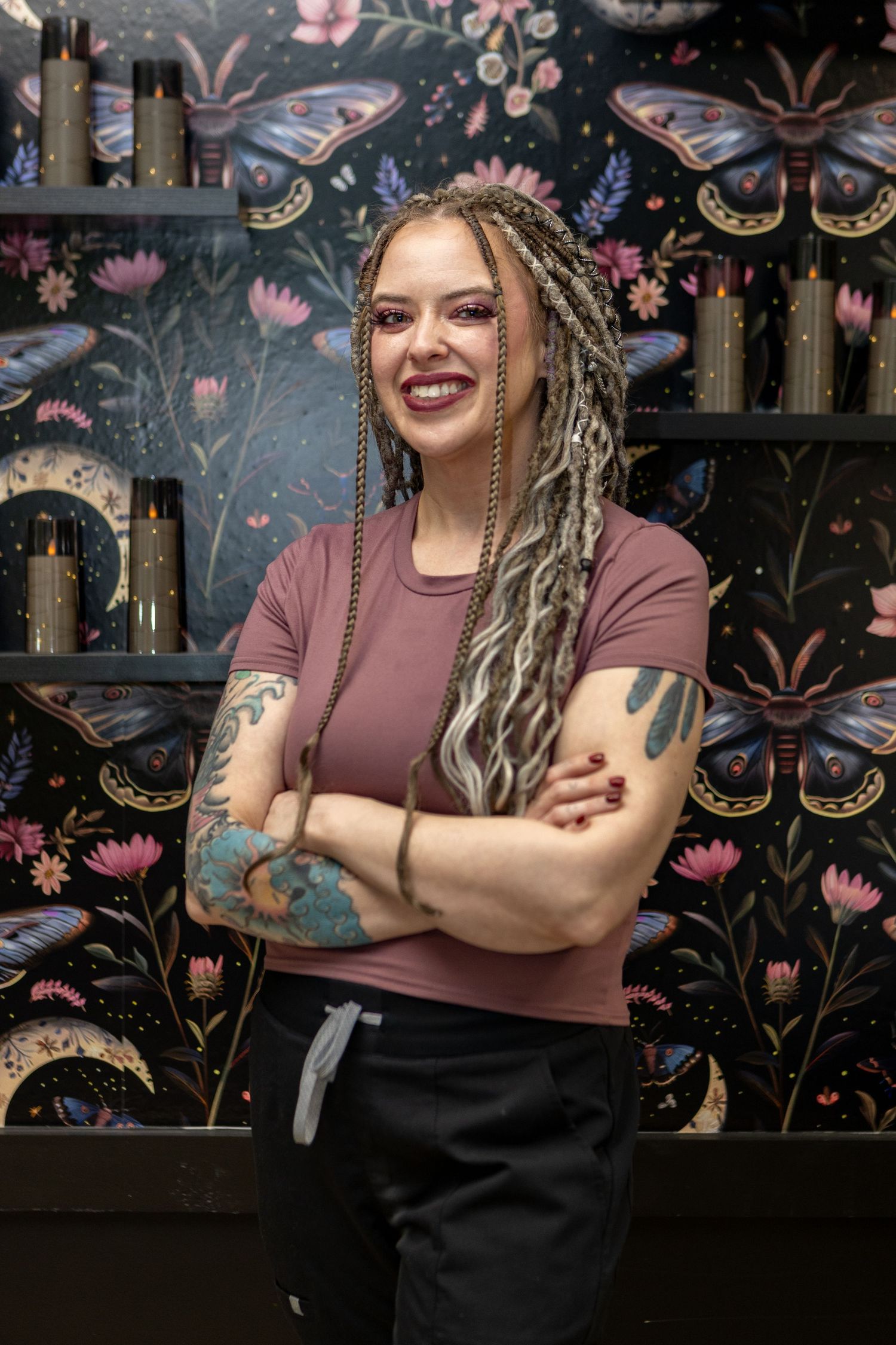 Tattooed woman with long braided dreads smiling with arms crossed in a salon with butterfly wallpaper.
