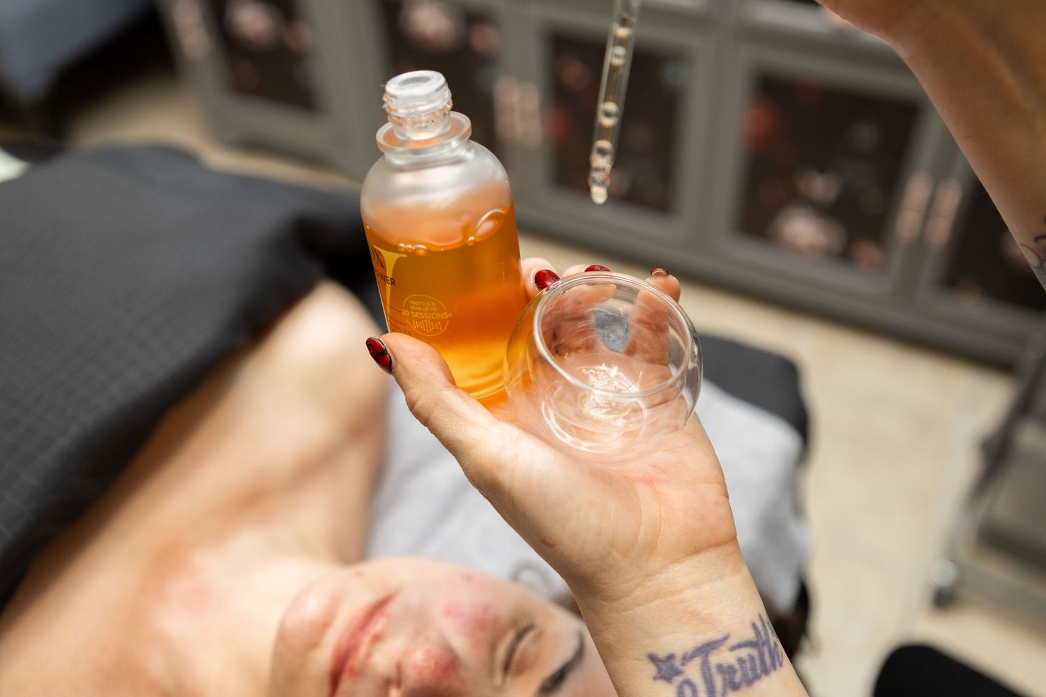 A tattooed hand pours amber facial oil into a glass bowl during a skincare treatment session.