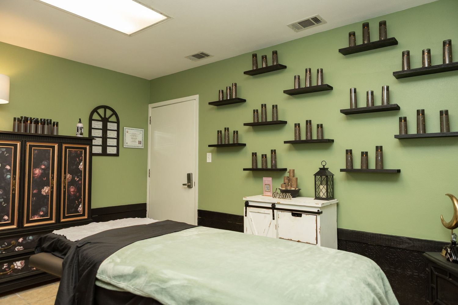 A serene spa treatment room with green walls, shelved candles, and a massage table with teal linens.
