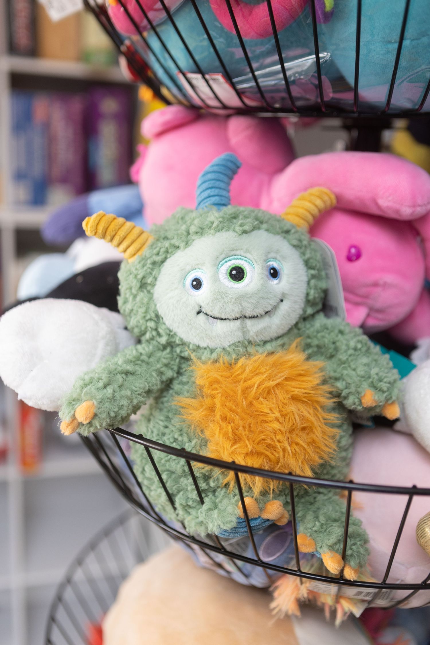 Cute green monster stuffed plush toy smiling in a wire basket surrounded by colorful soft toys.
