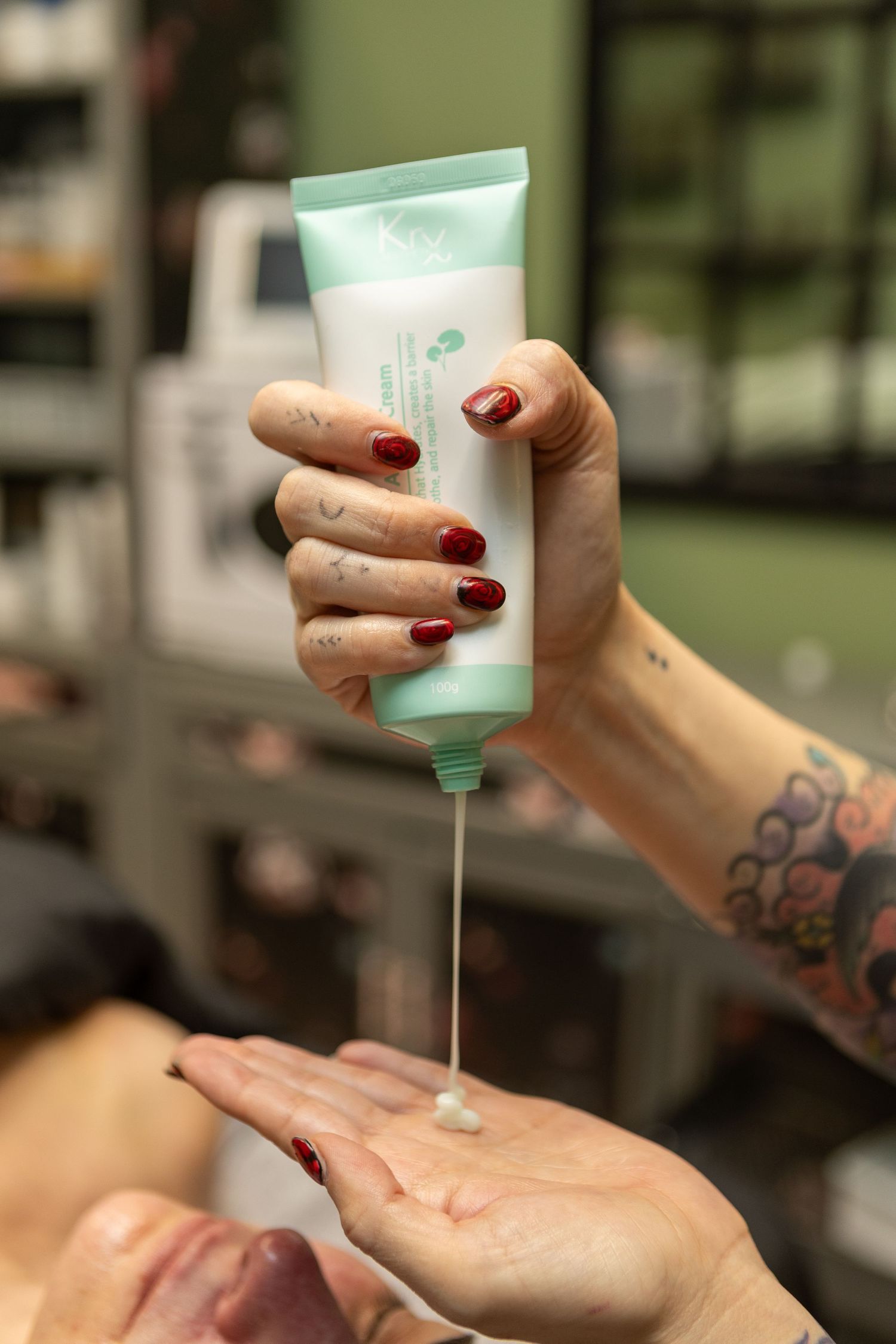 Tattooed hand with red nails squeezing mint green skincare tube, dispensing cream onto open palm in beauty salon.