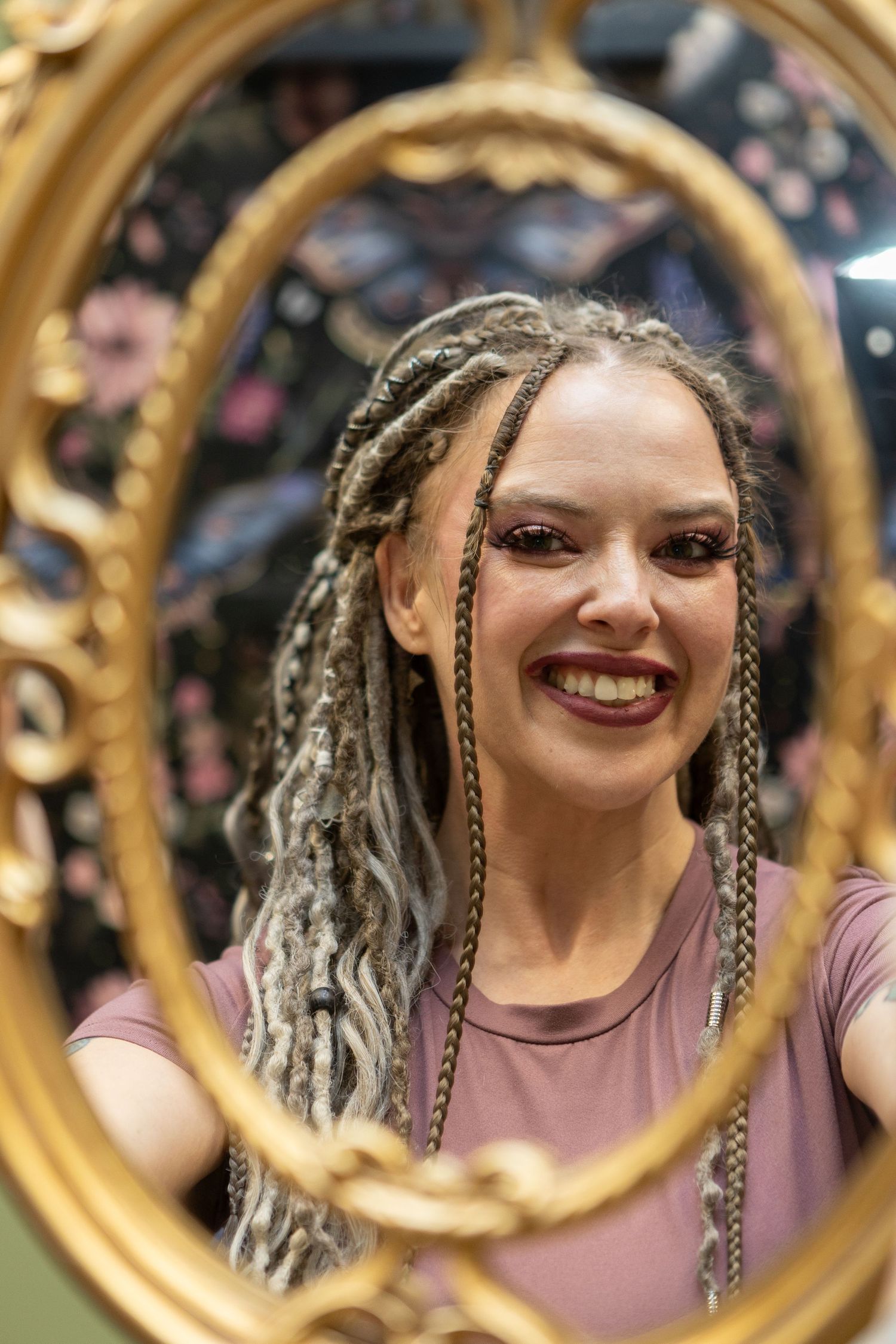 Woman with braided dreadlocks smiling at her reflection in an ornate gold oval mirror in a salon.