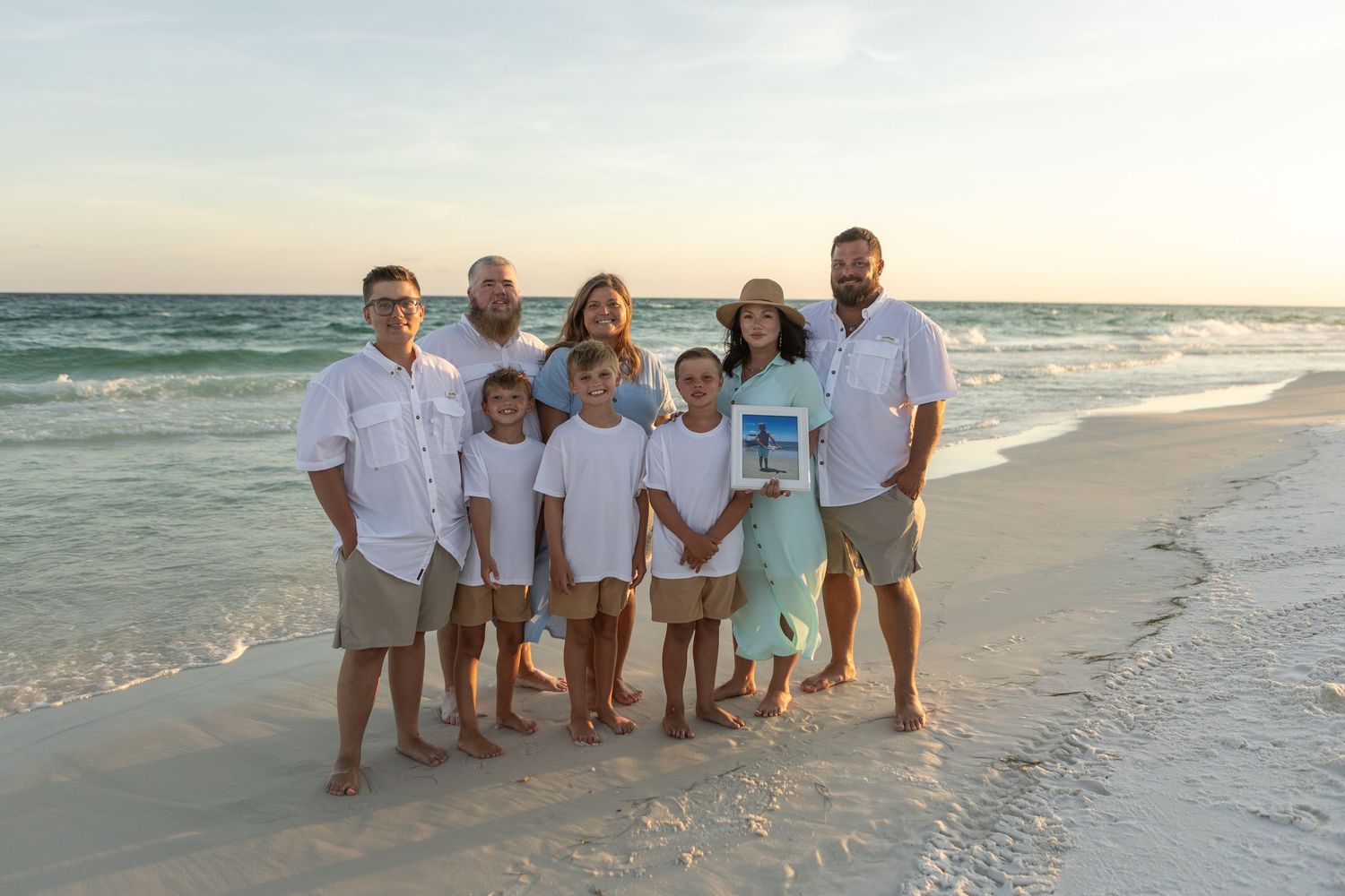 Extended family group poses on a sunny beach holding a framed photo with clear turquoise water behind them.