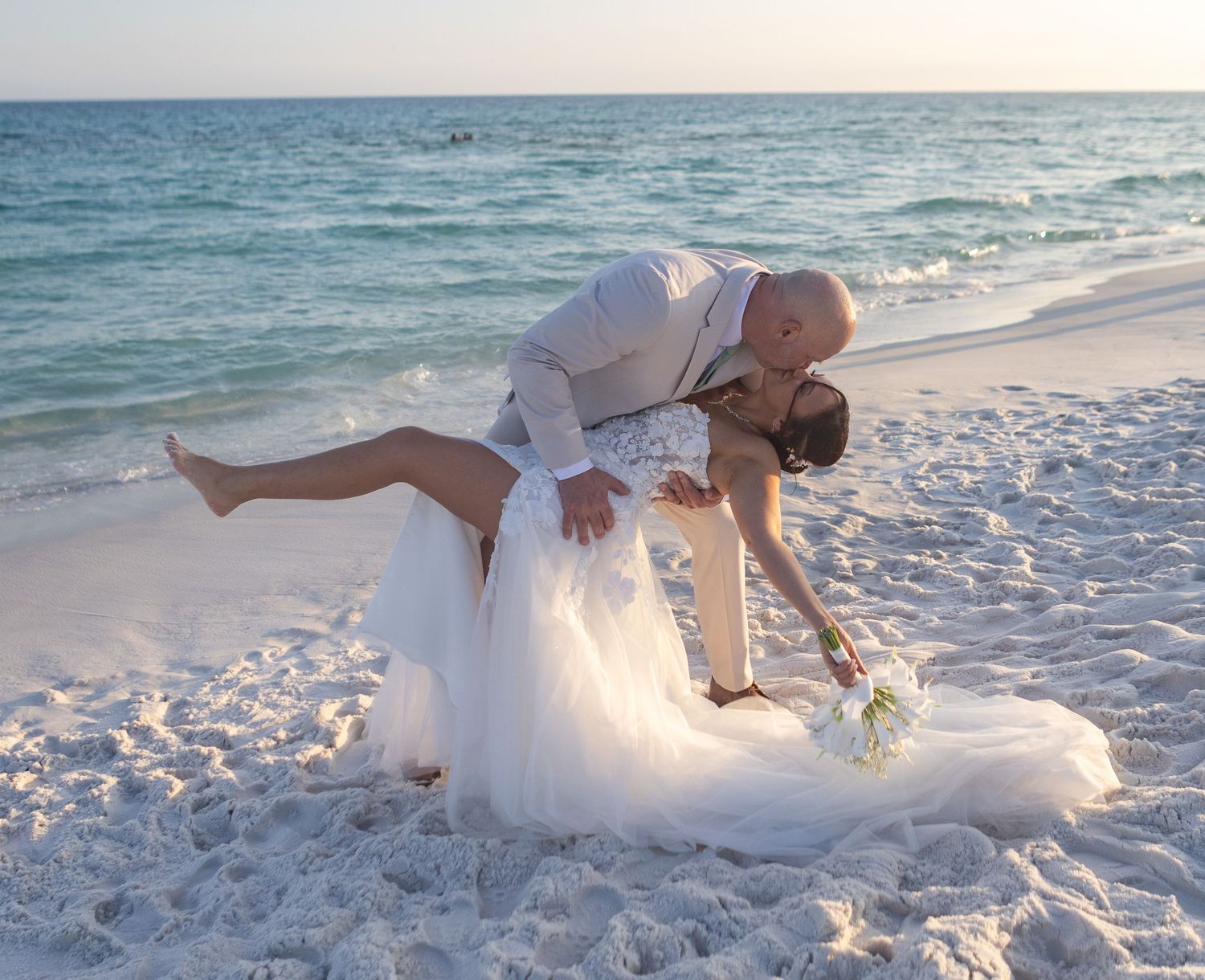 Bride and groom share a romantic dip kiss on a white sand beach with turquoise ocean waves in the background.