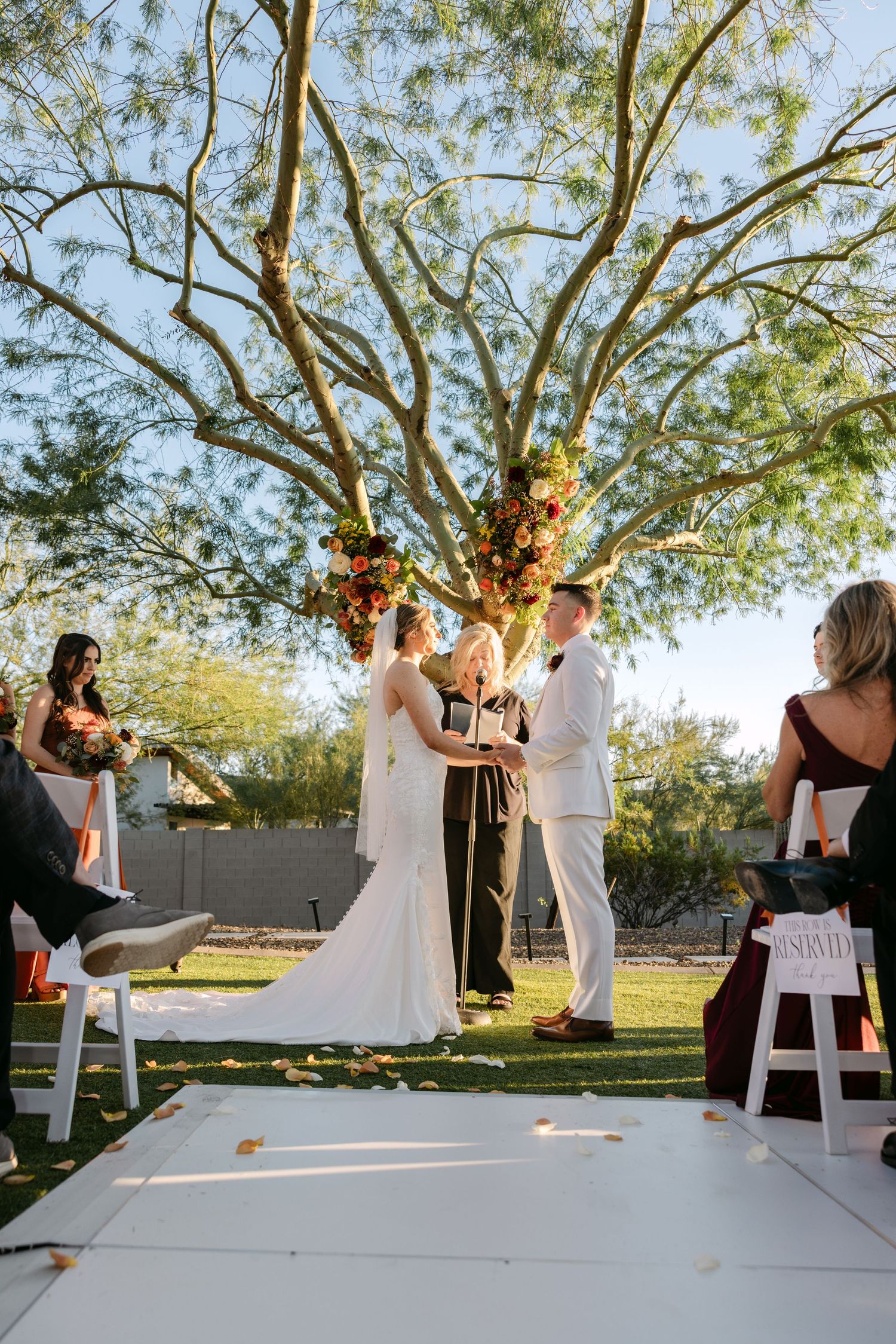 A wedding ceremony takes place under a large tree with guests watching the couple exchange vows in an outdoor setting.