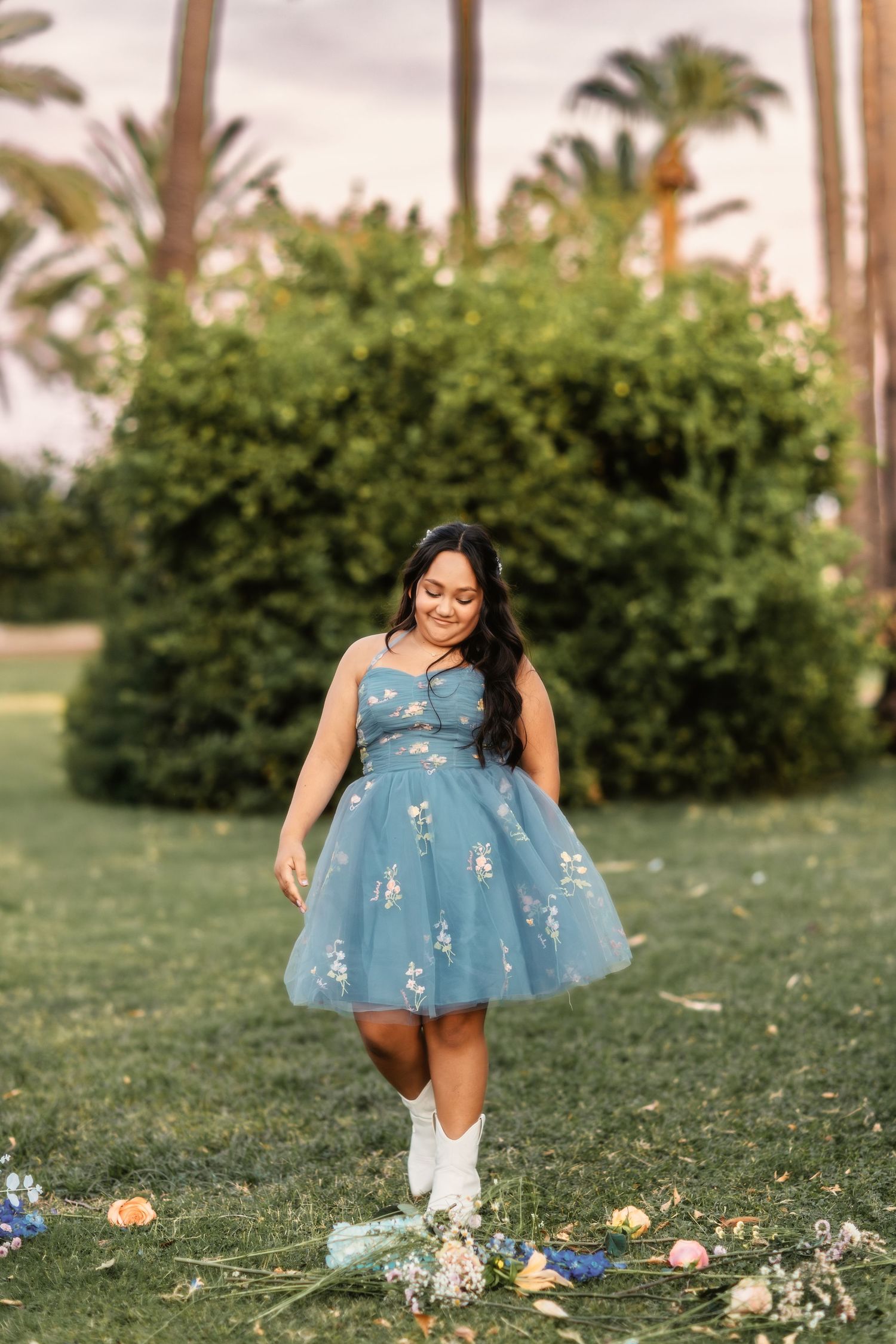 A person in a light blue tulle dress with floral details walks through a garden setting with palm trees in the background.