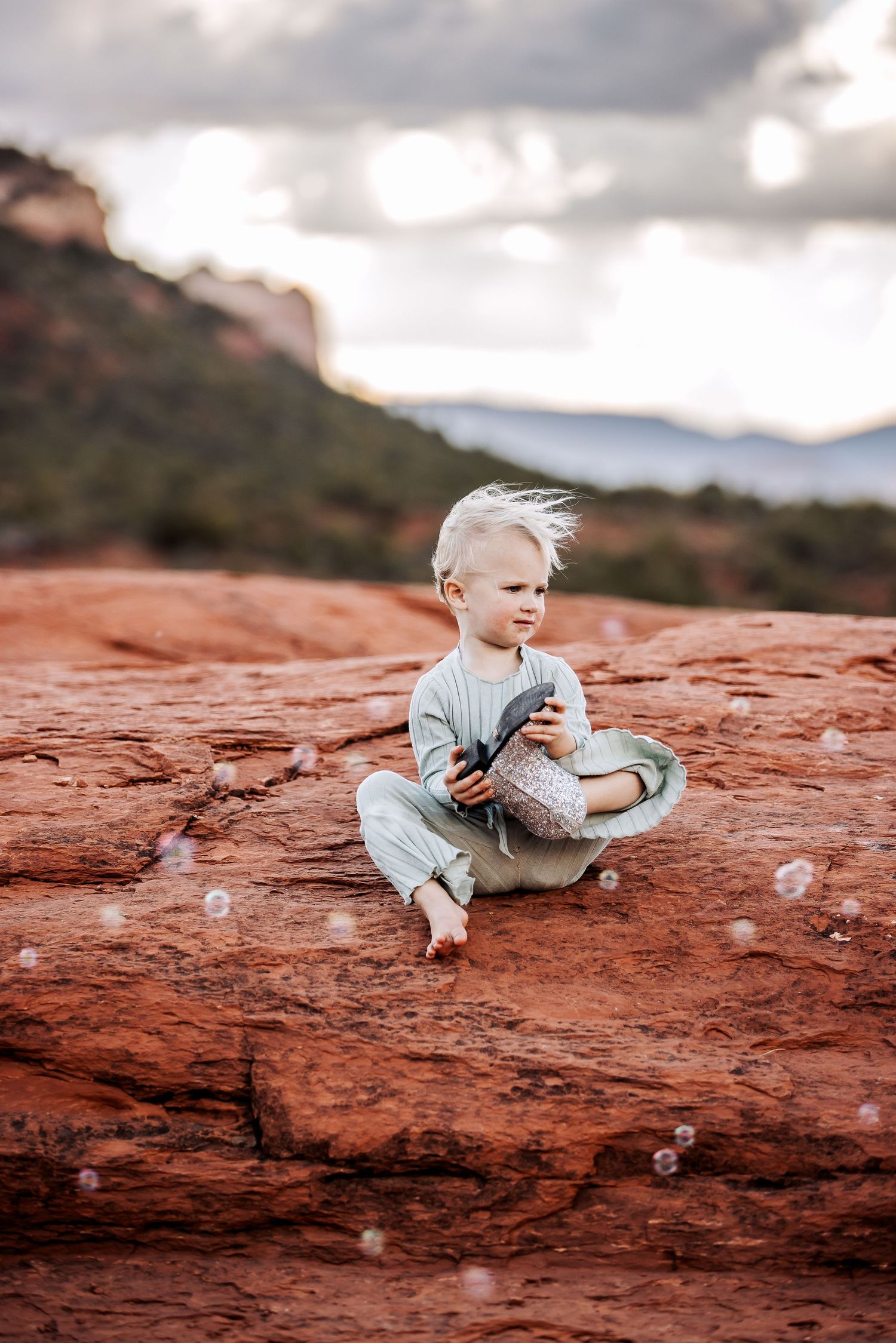 Small child in light blue outfit sits on vibrant red rocks in a desert landscape with mountains in background.