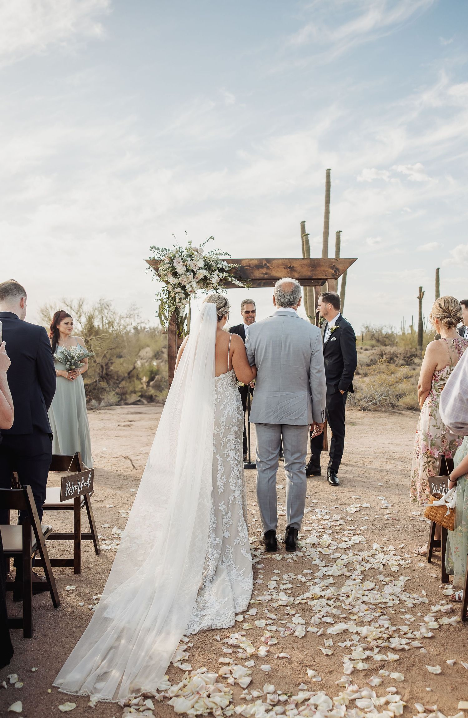 Wedding ceremony setup in a desert landscape with scattered rose petals leading to wooden chairs and an altar.