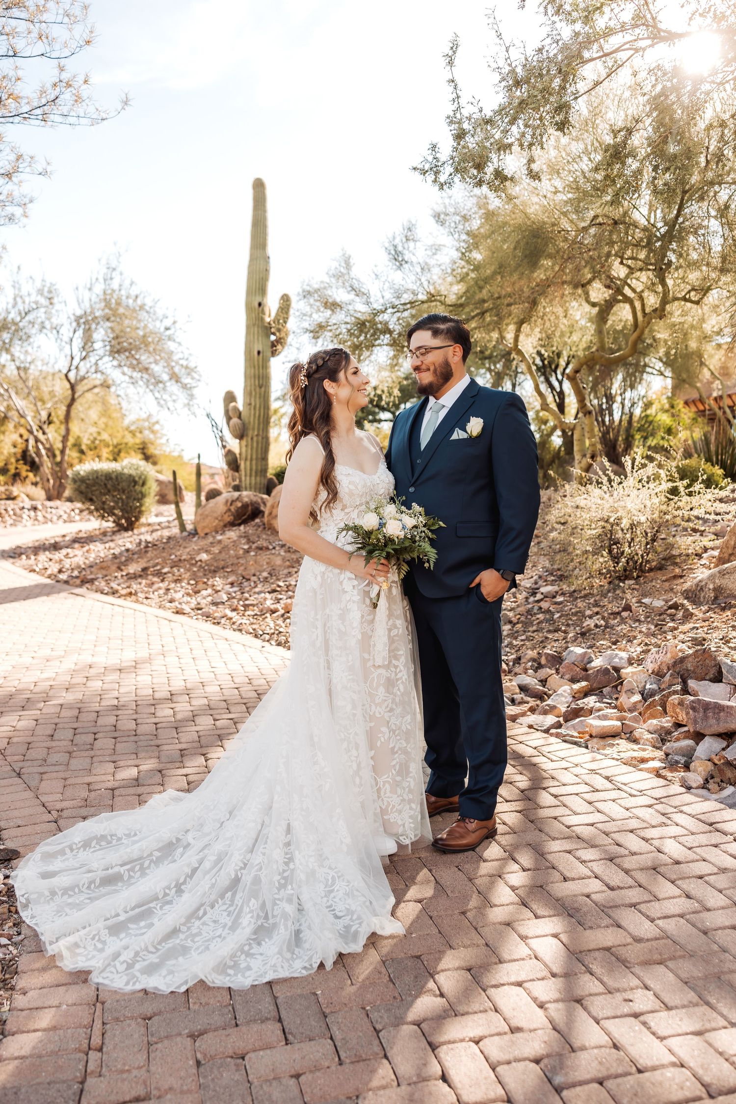 Wedding couple poses together on brick pathway with desert cacti and sunlit landscape in background.
