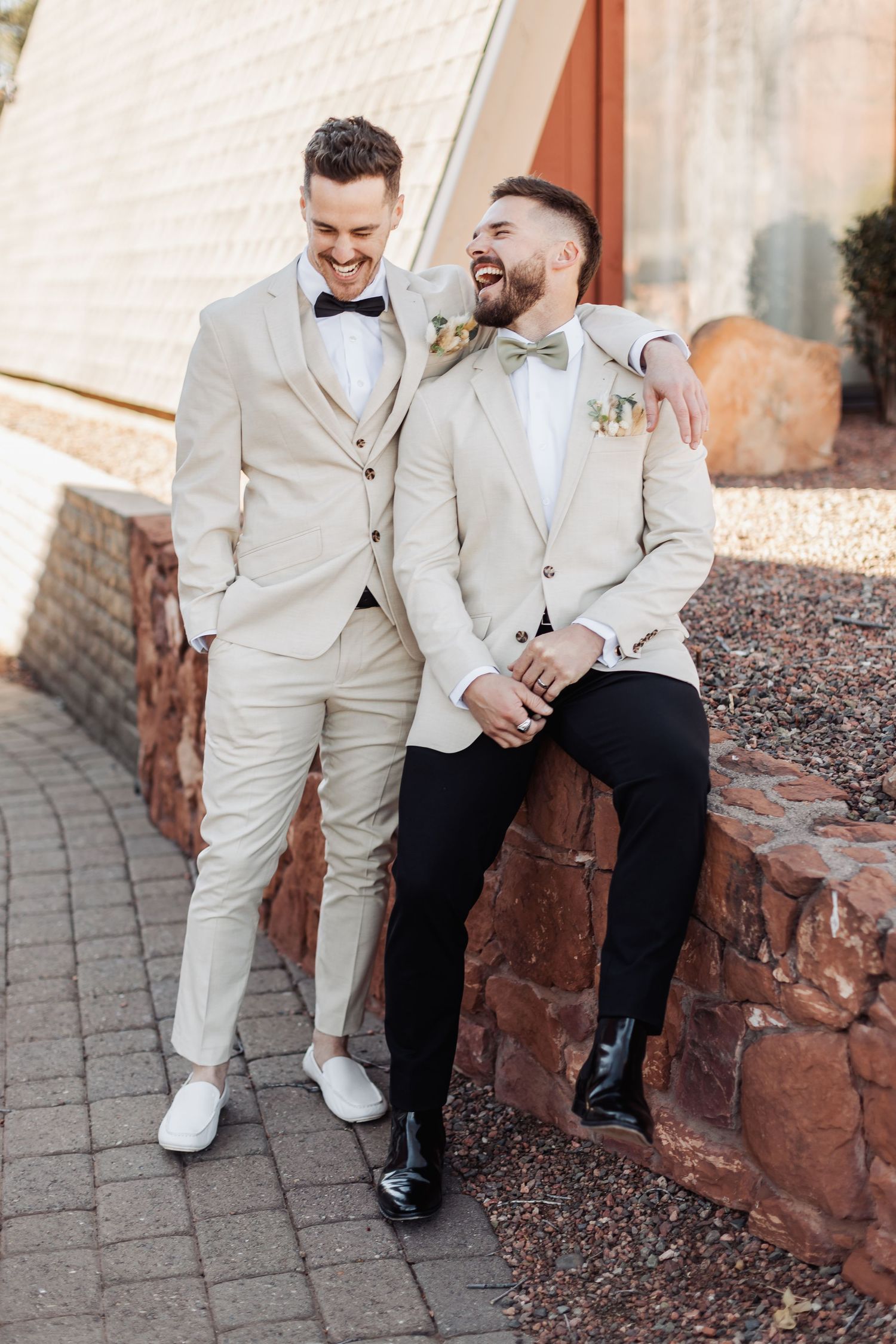 Wedding party members in light beige suits and dark attire sitting casually on stone wall.