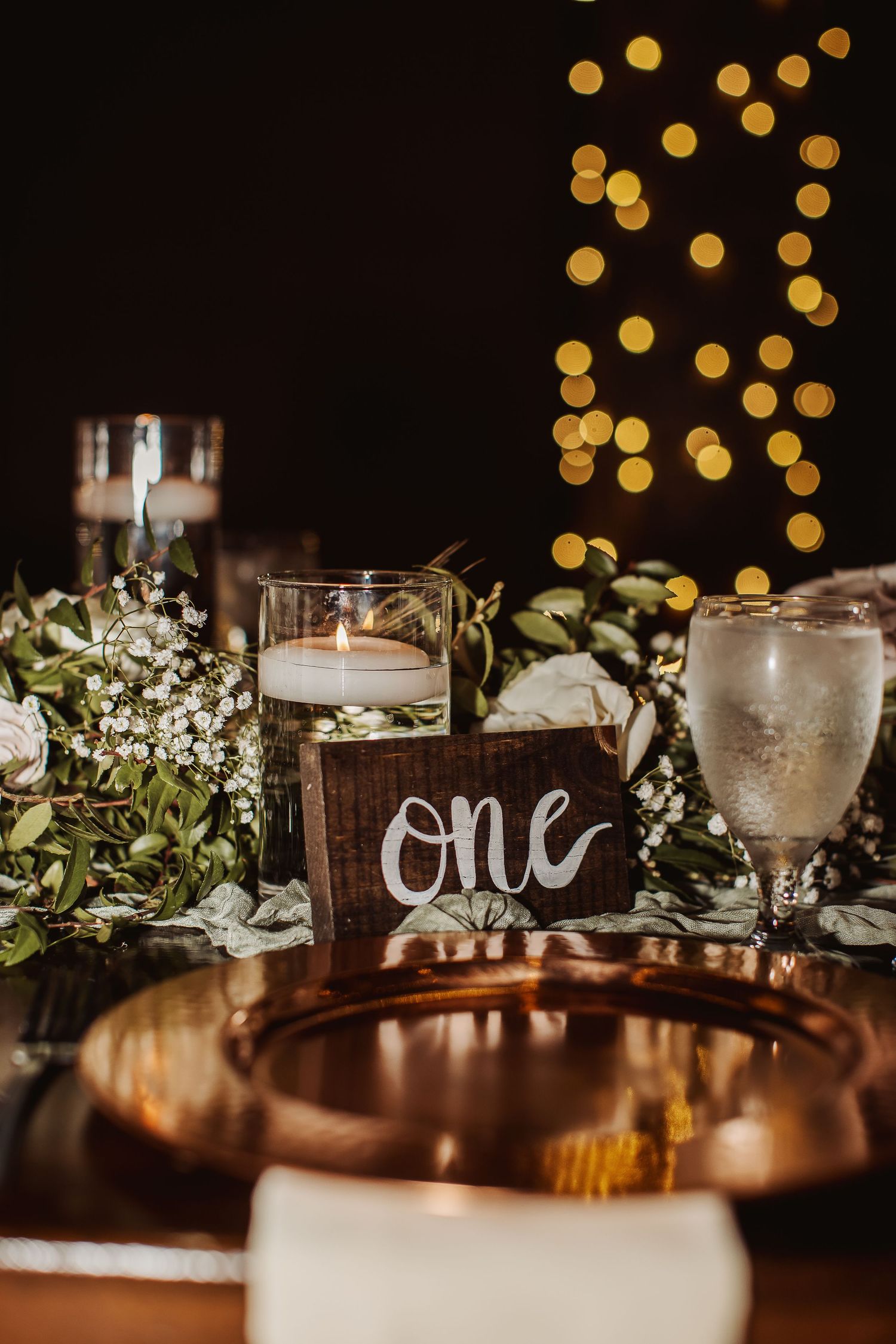 Wedding reception table decor with rustic wooden signs, glowing bokeh lights, and delicate floral arrangements.