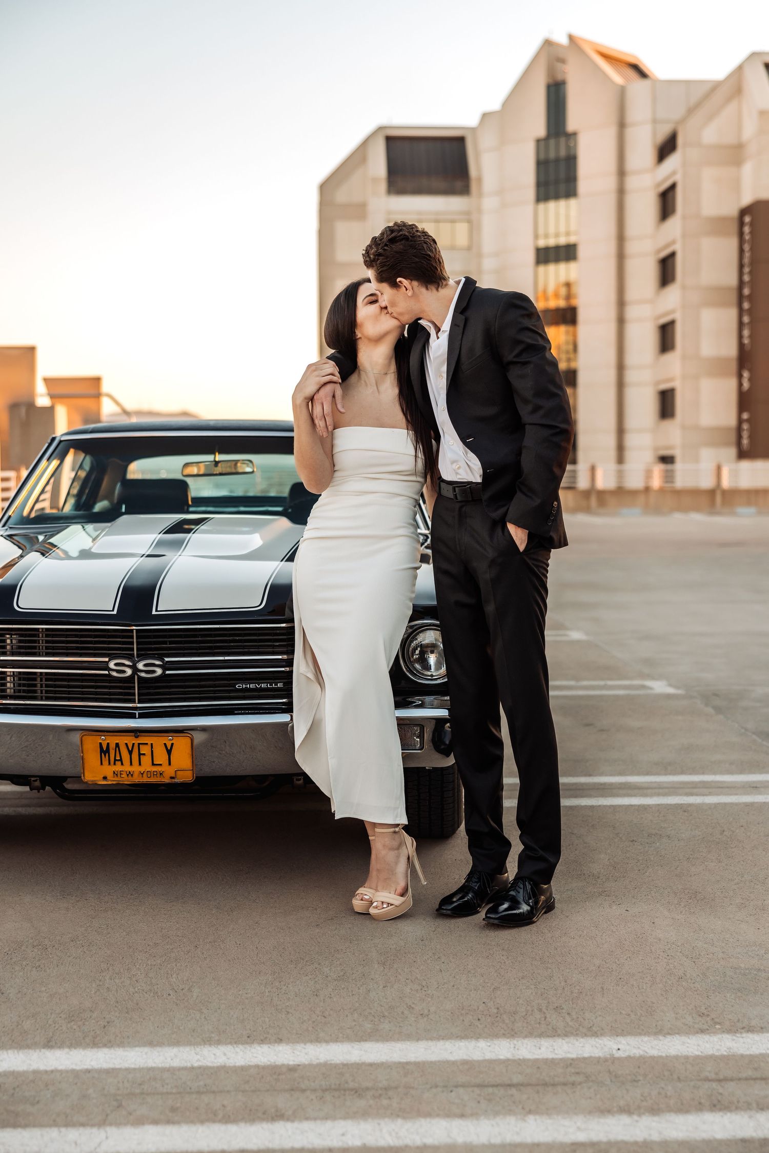 Romantic couple poses together in wedding attire next to a classic Chevrolet SS car during golden hour.