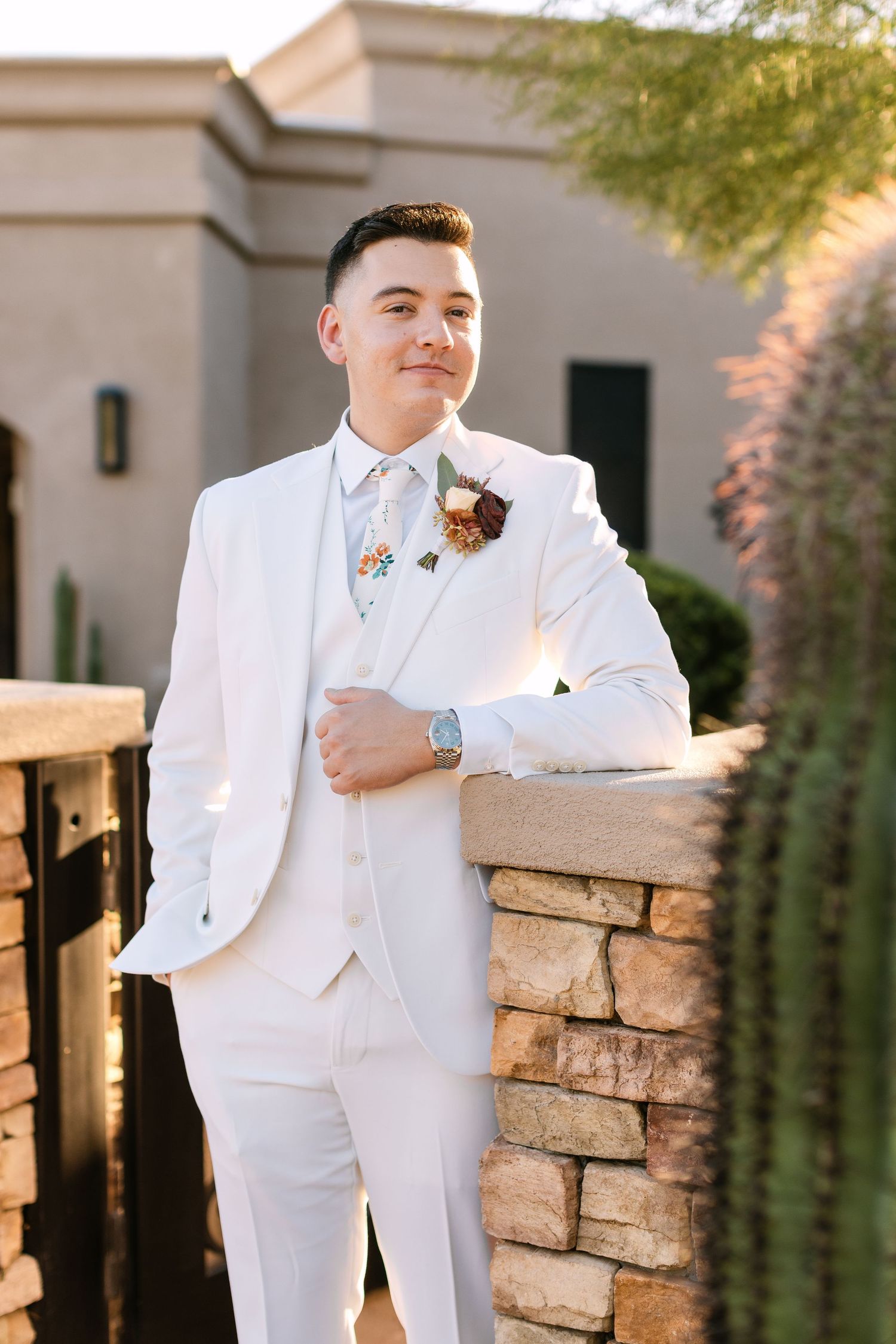 Someone wearing a crisp white wedding suit with boutonniere poses confidently against a stone wall in warm sunlight.