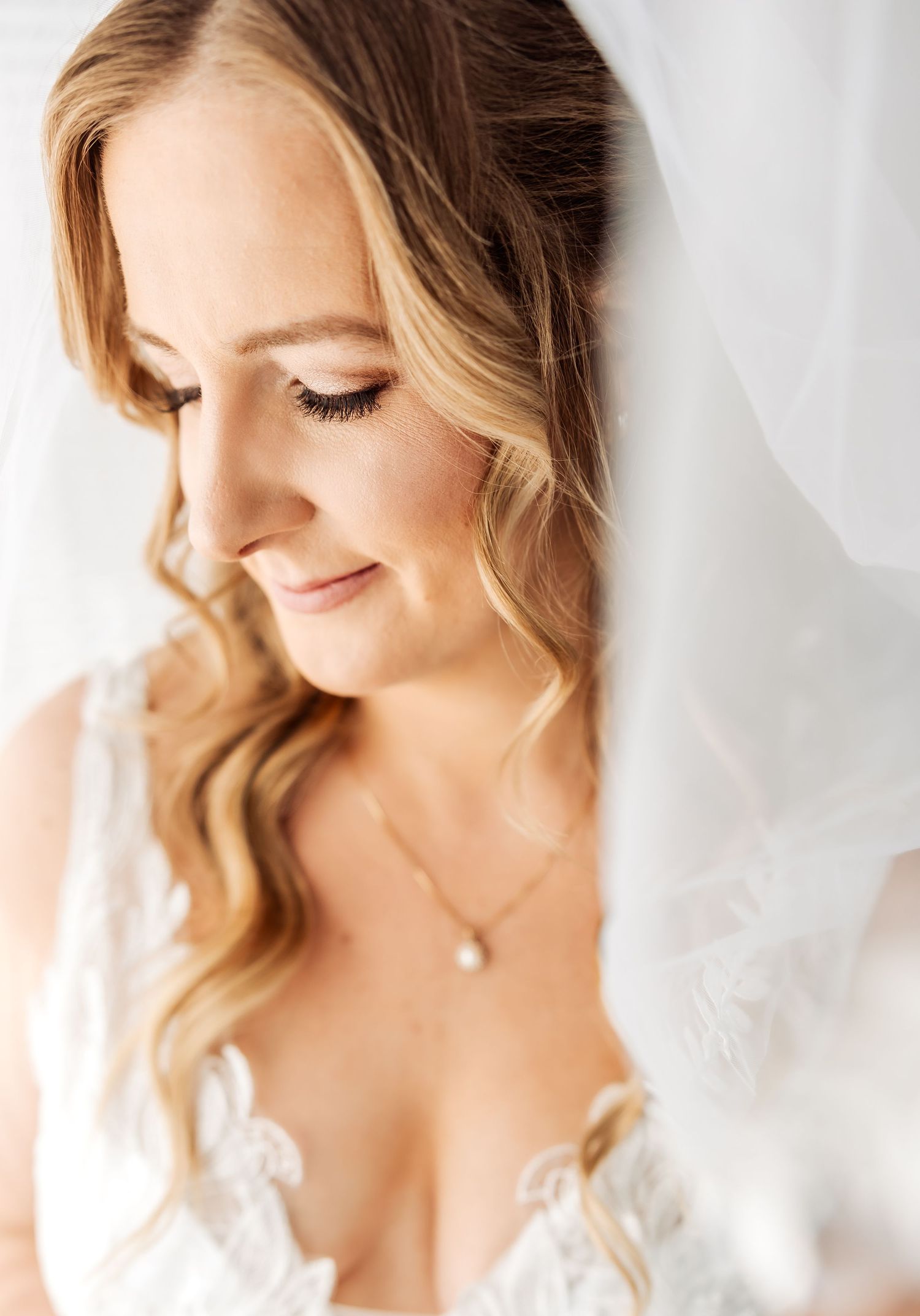 Close-up portrait of a bride with soft makeup and wavy blonde hair wearing pearl earrings against a white background.