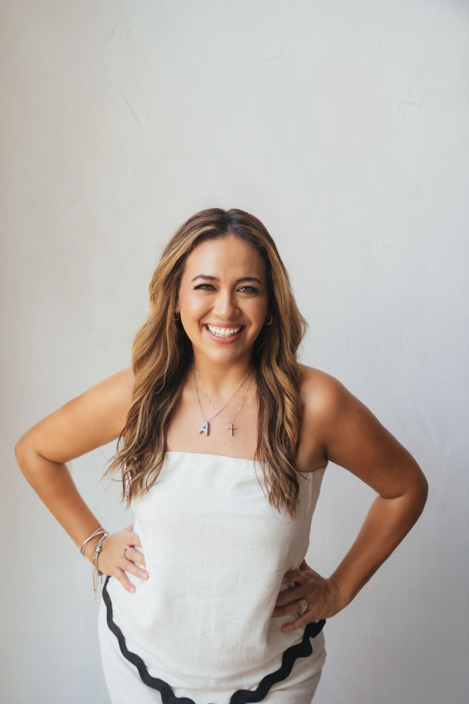 A woman with wavy brown hair wearing a white sleeveless top poses confidently against a light gray background.
