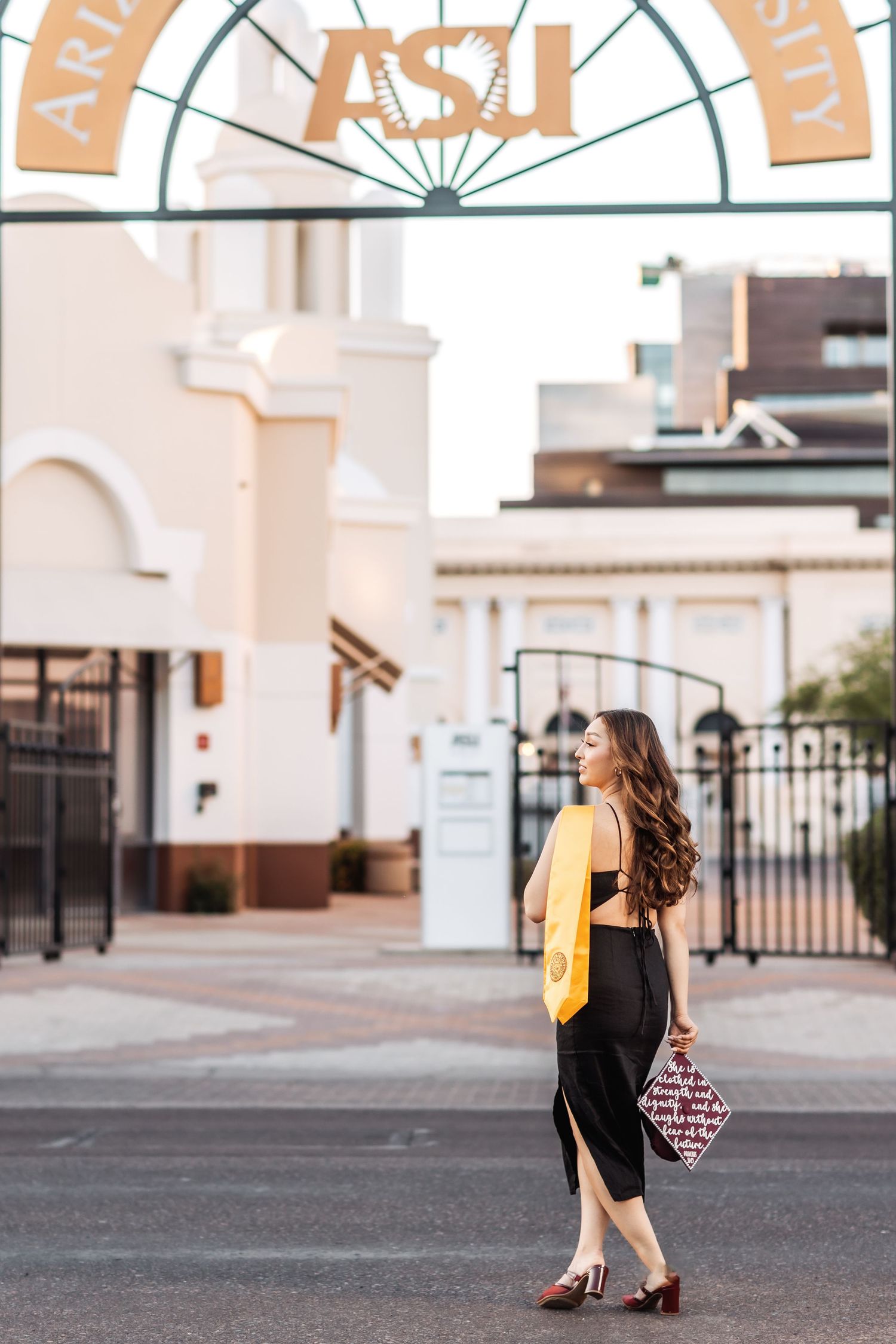 Elegant graduation photo series of a student in a black dress and gold backpack walking away from a white building archway.