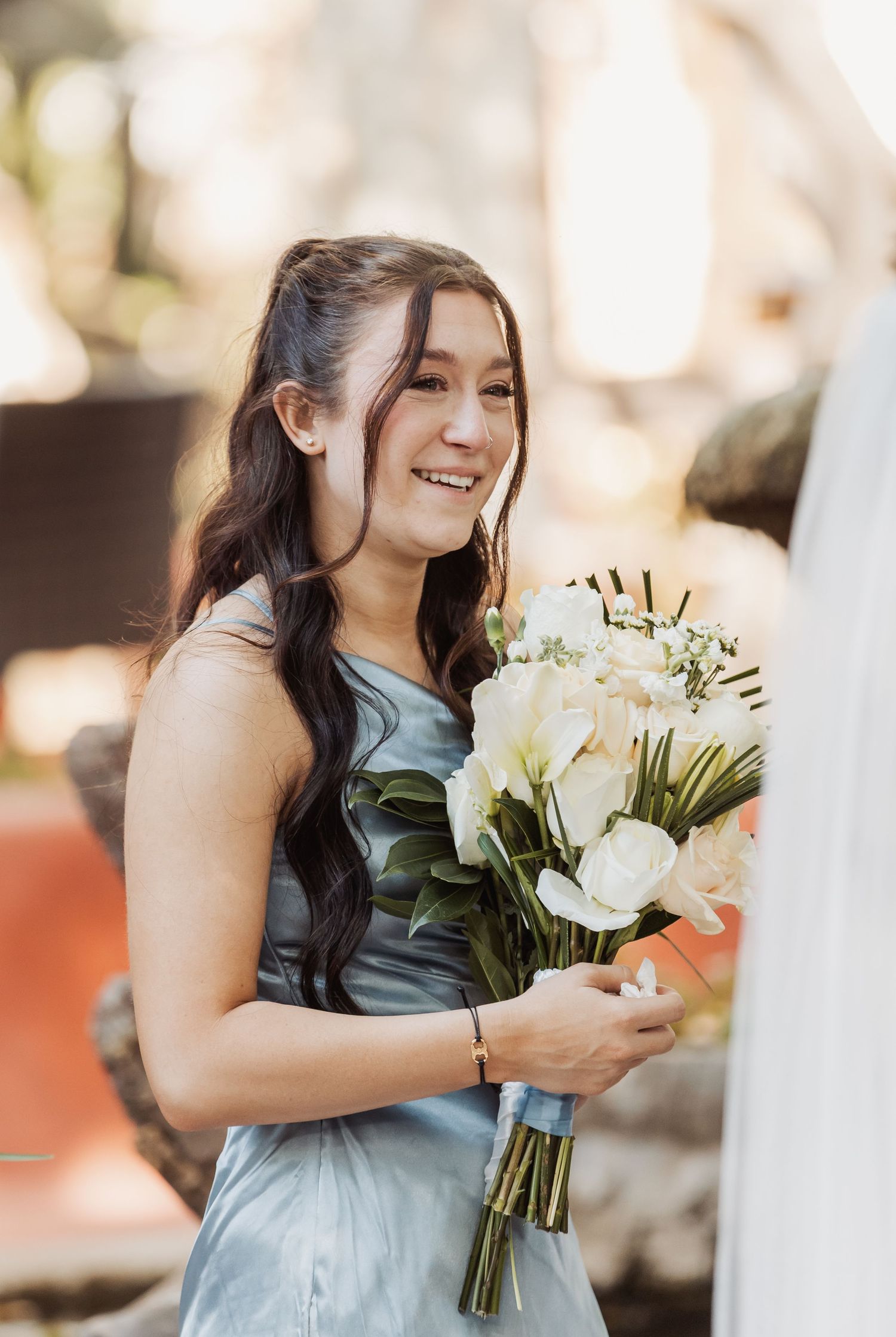 Bridesmaid in a light blue dress holds a white floral bouquet while smiling during outdoor wedding photos.