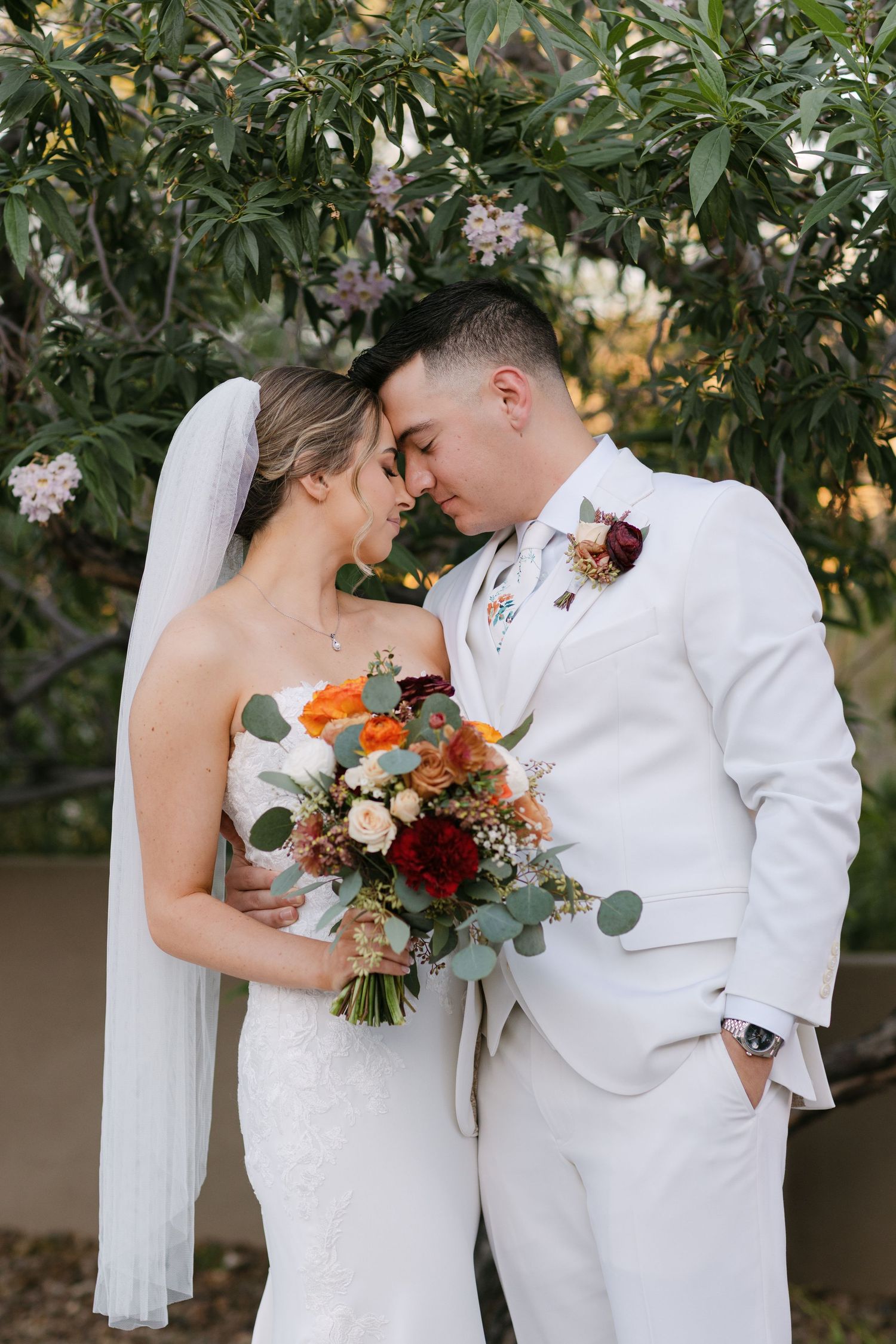 Romantic wedding portraits featuring a couple in white outfits with a colorful autumn bouquet against greenery.
