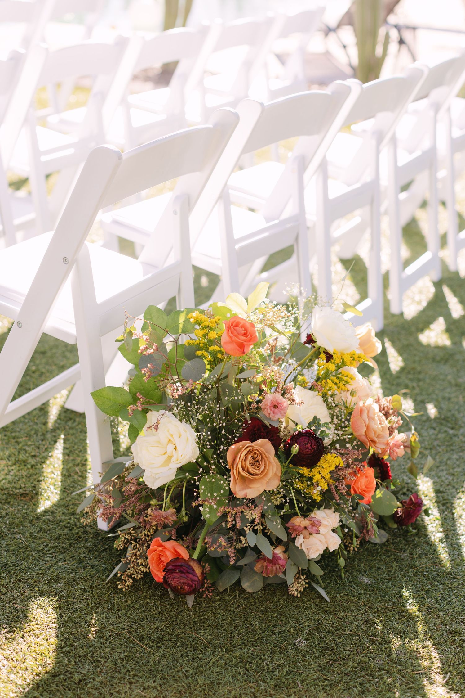 Beautiful floral arrangements with coral and burgundy blooms decorating white ceremony chairs on grass.