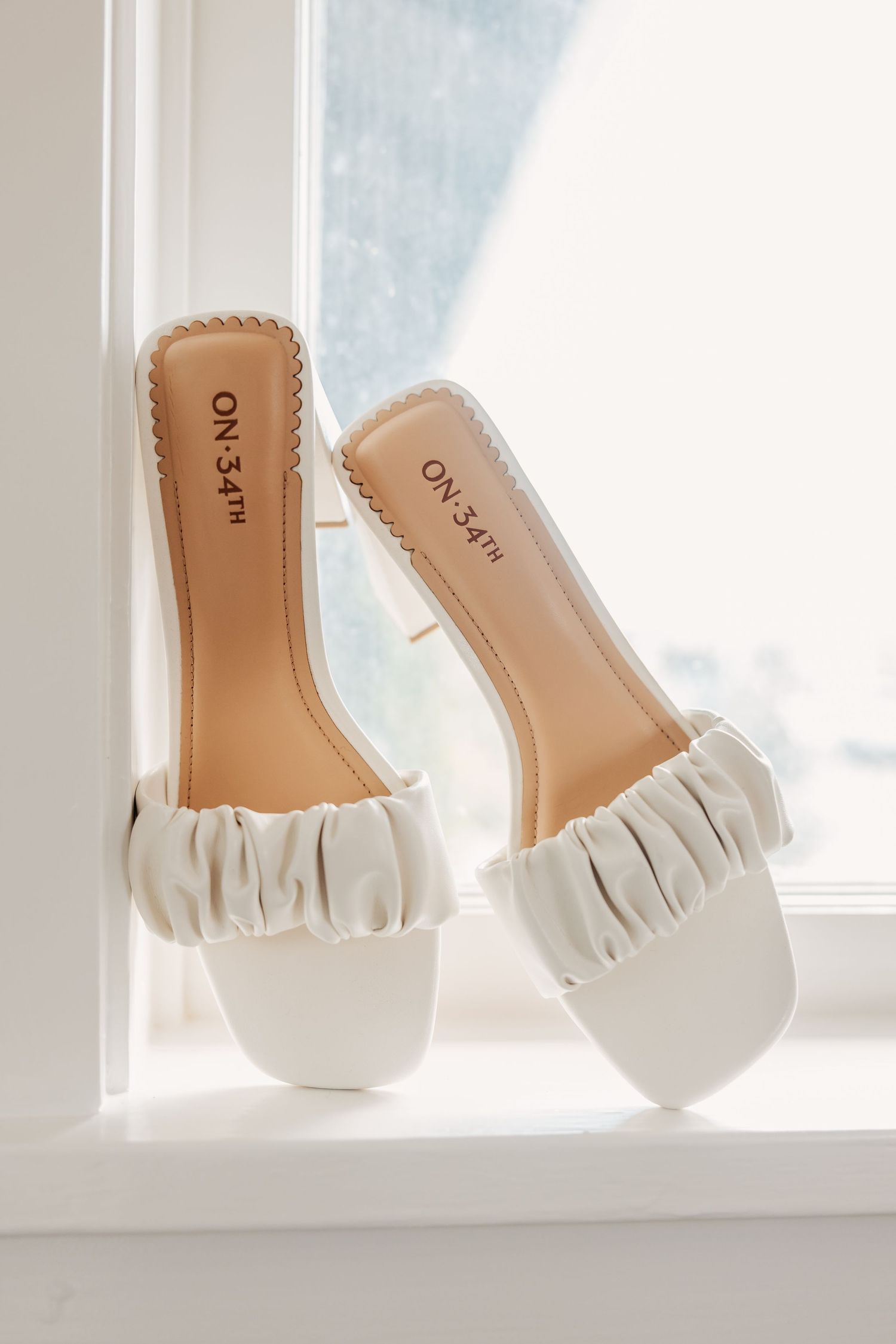 White leather bridal flats with ruffled details displayed on a windowsill in natural light.