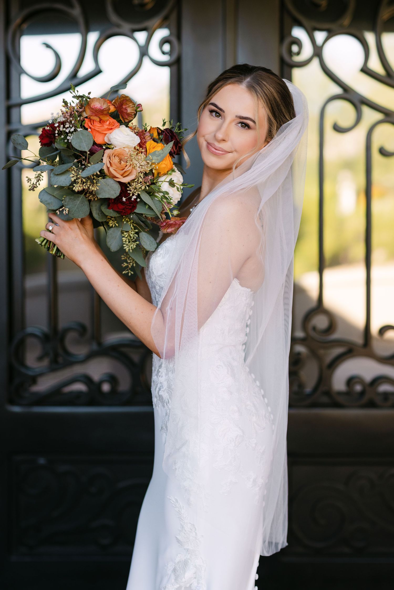 A bride in a white wedding dress and veil poses with her peach and coral bouquet against an ornate iron gate.