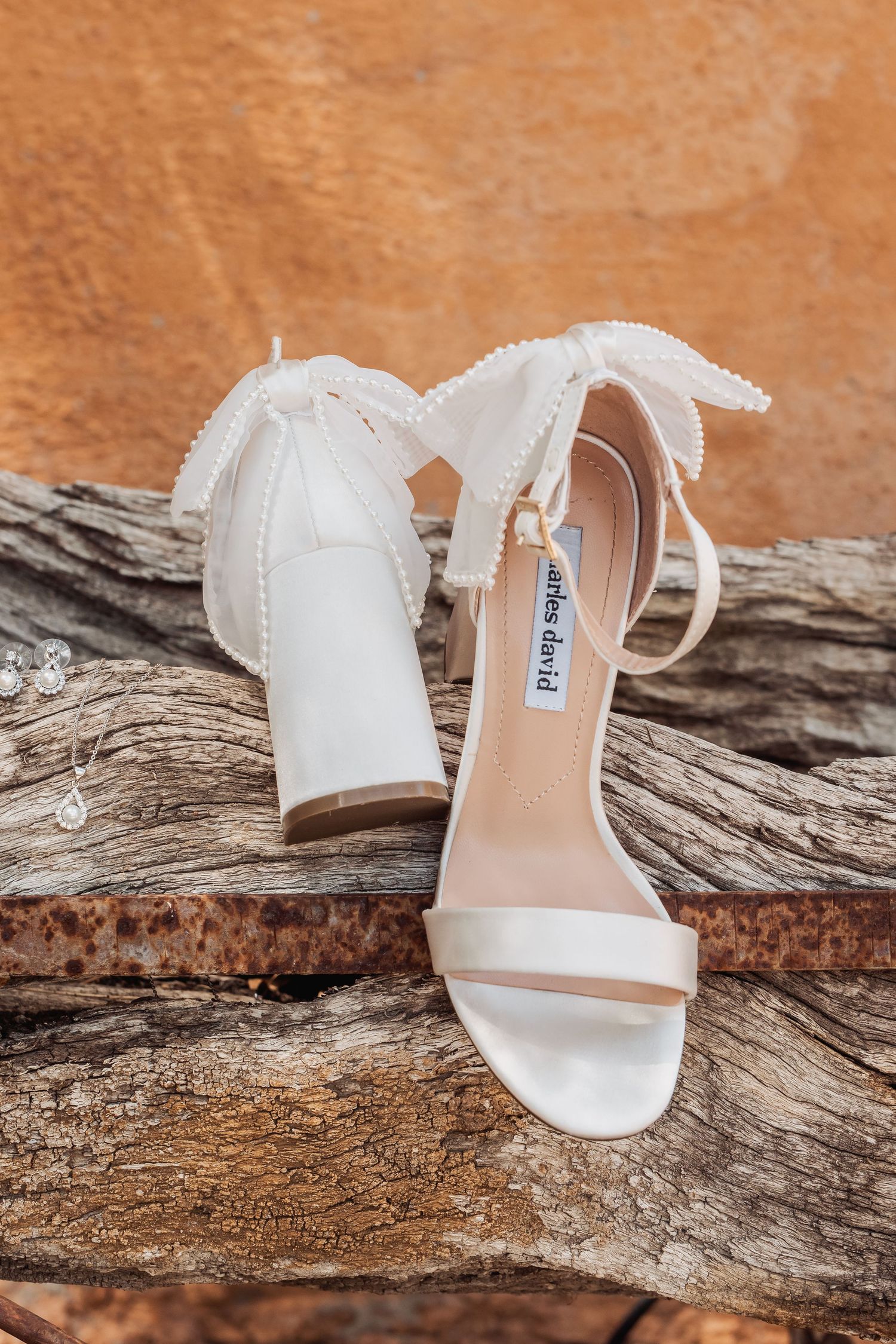White bridal sandals with ankle straps displayed on weathered driftwood against a rustic brown background.