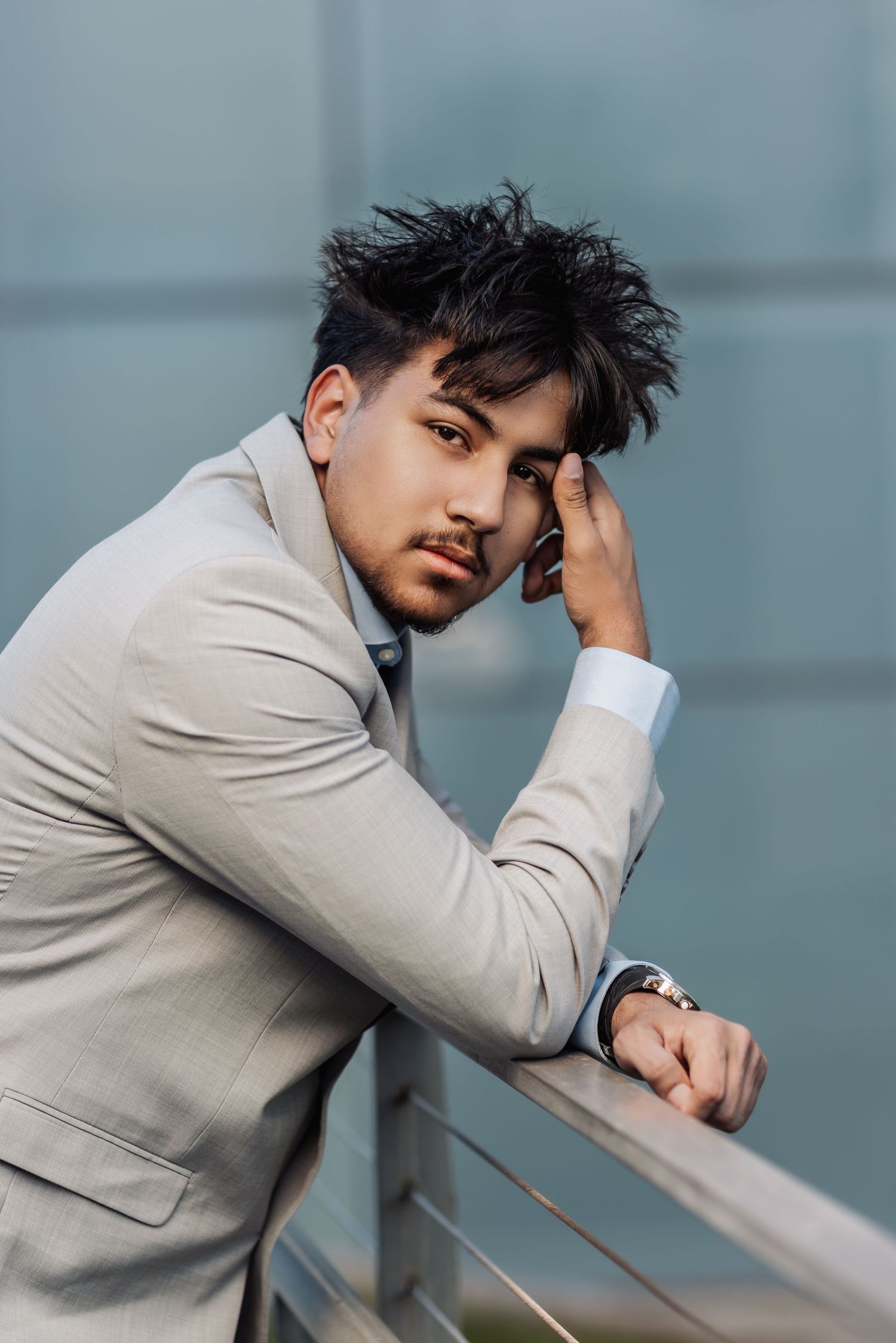 A person in a light gray suit jacket poses thoughtfully against a railing with a modern blurred background.