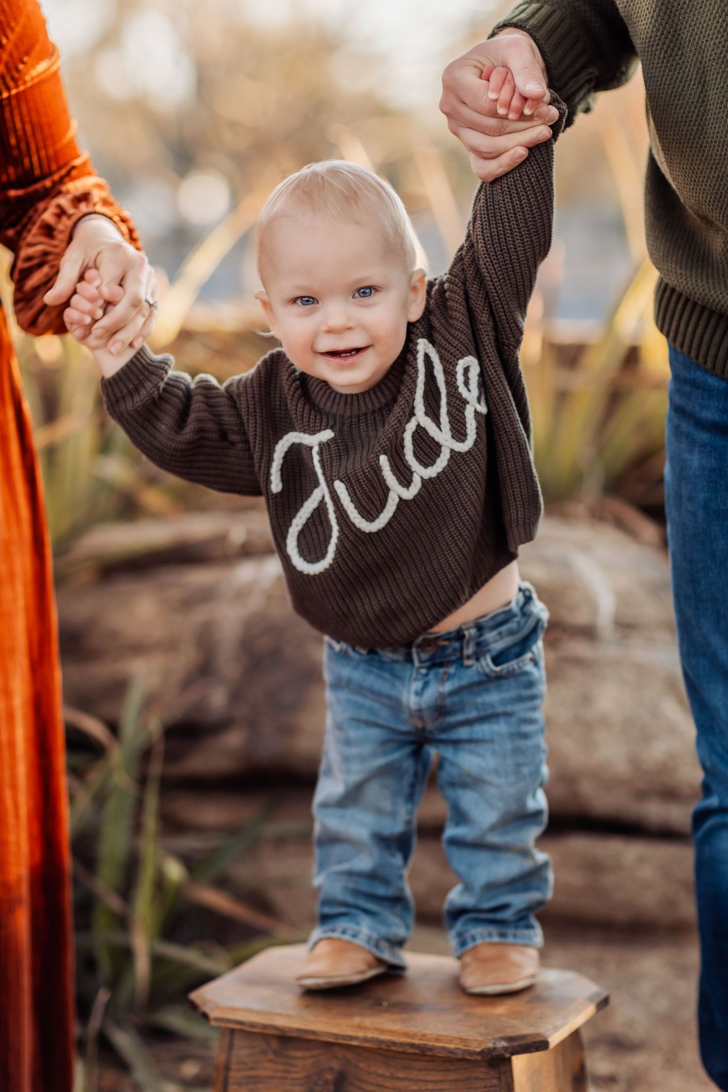 Toddler in brown sweater and jeans stands on wooden block holding hands with parents in autumn outdoor setting.
