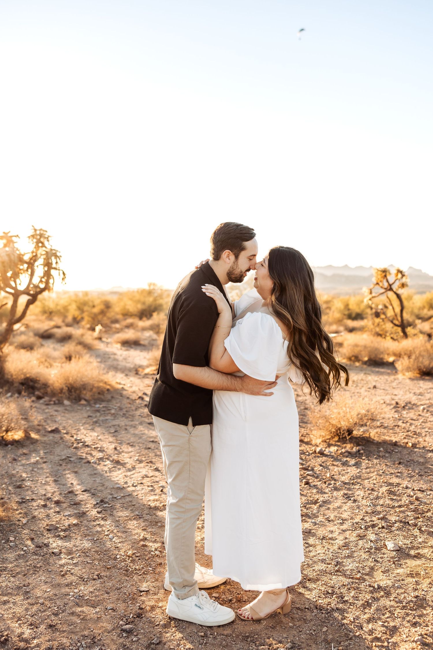 Couple embraces in the desert at golden hour with Joshua trees in the background and soft sunlight illuminating the scene.