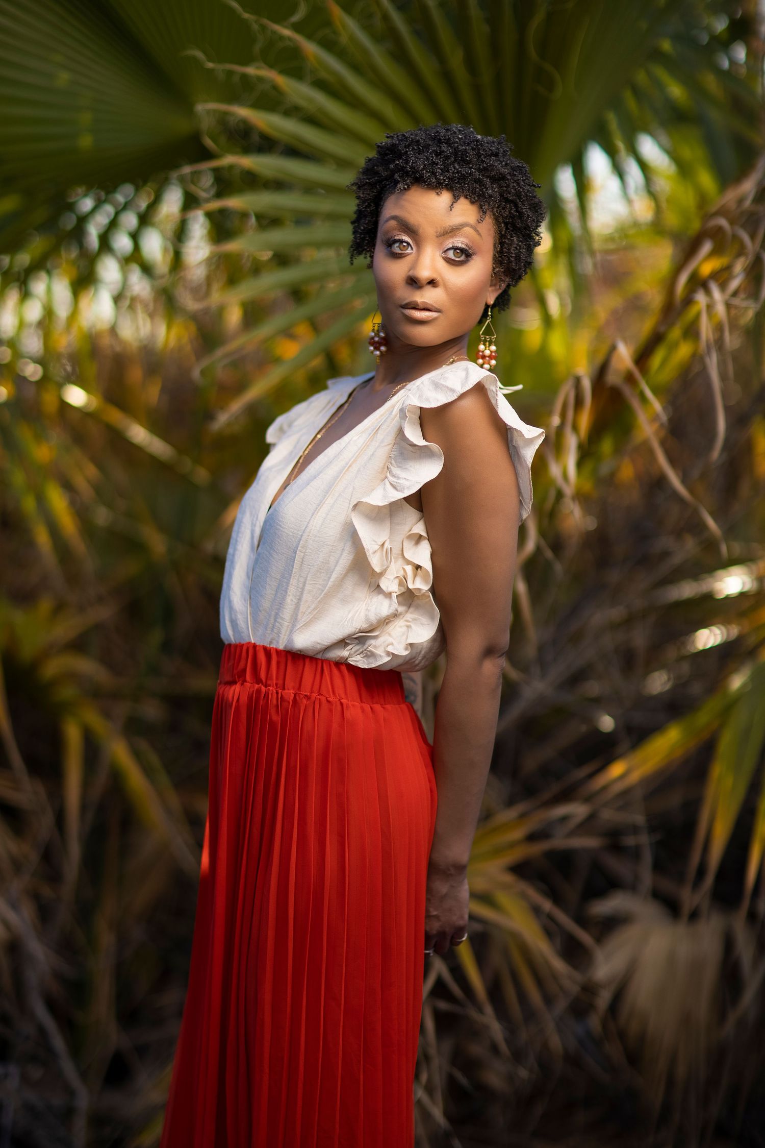 A person in a ruffled white blouse and flowing red maxi skirt poses against a tropical palm backdrop in golden light.