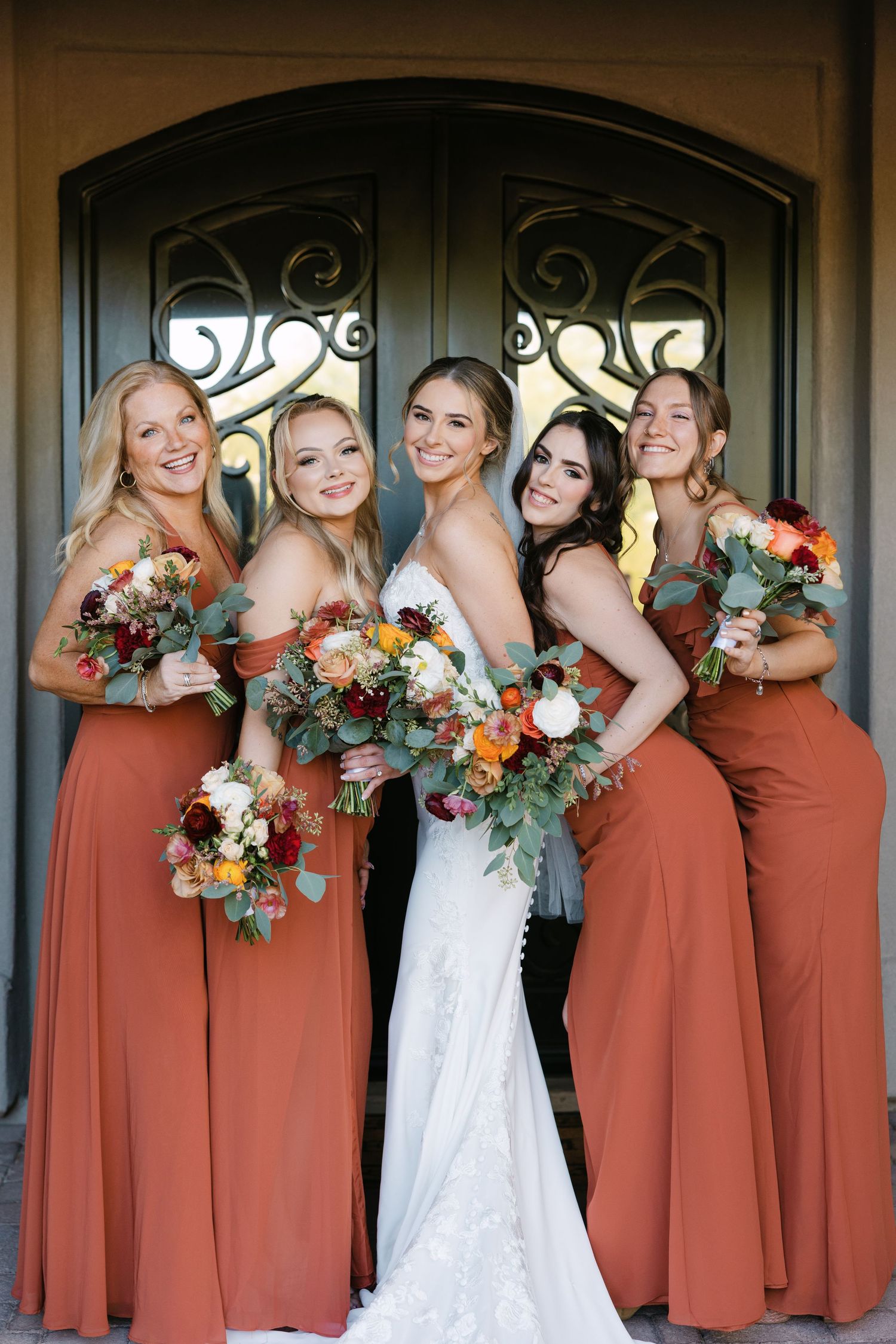 Bridesmaids in matching terracotta dresses pose with bride in white gown holding autumn-colored bouquets.
