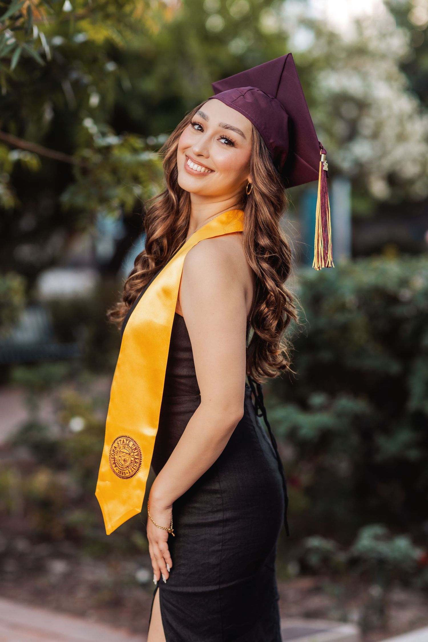 Graduate in black dress and gold sash poses outdoors during graduation photo session against natural backdrop.