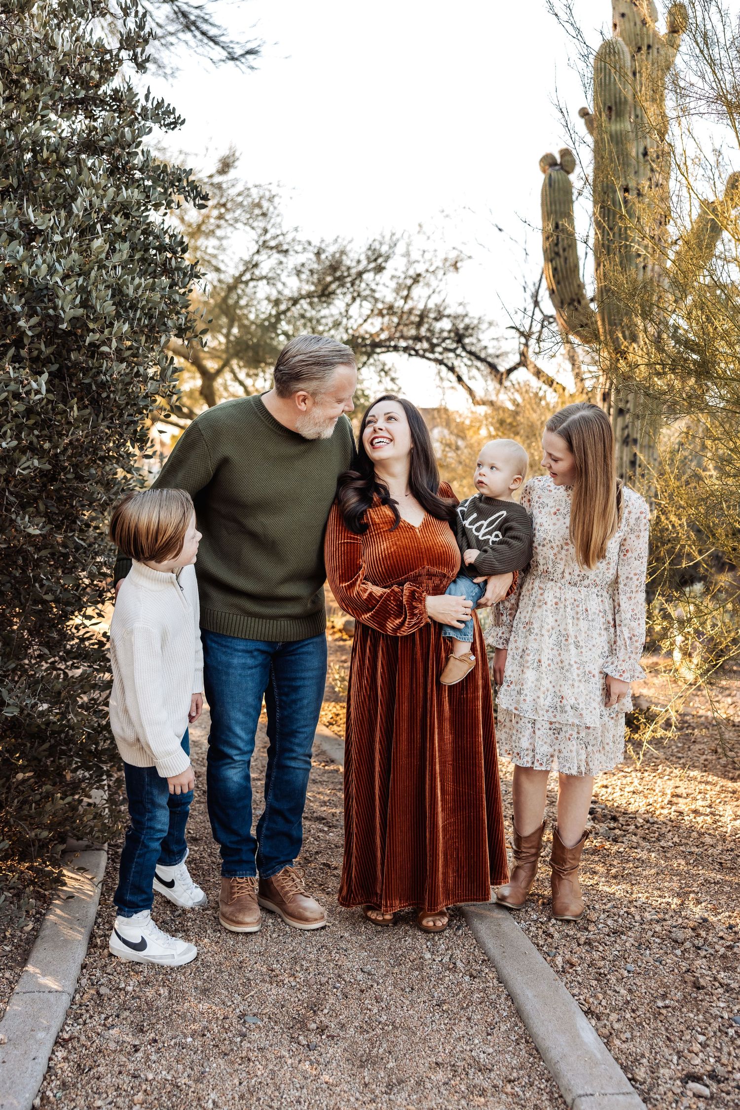 A candid family portrait shows playful interaction between family members on a desert trail at sunset.